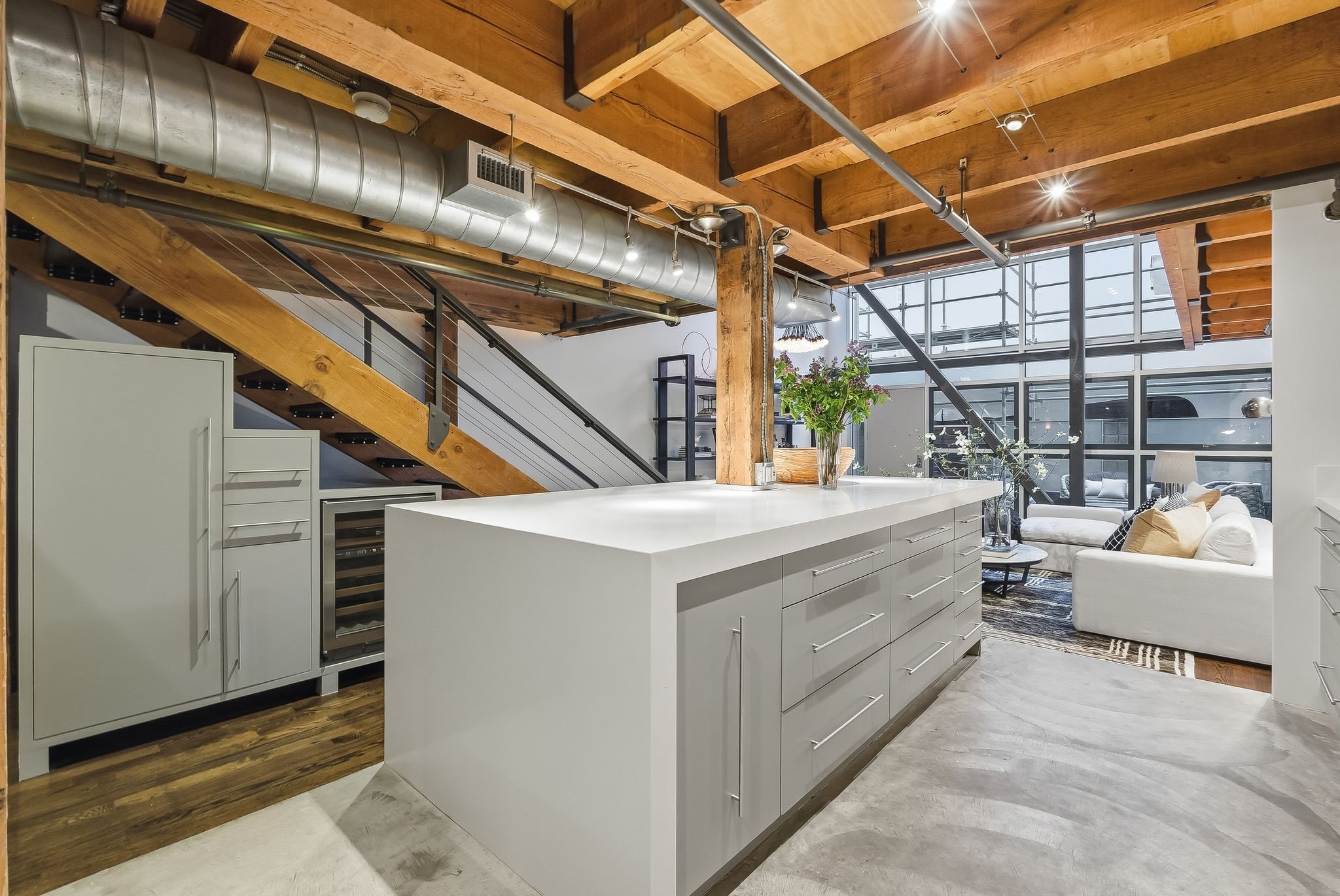 Modern open-concept kitchen with white cabinets and a large kitchen island, exposed wooden beams, industrial ductwork, and large windows with a view of a living area with a white sofa and a coffee table.