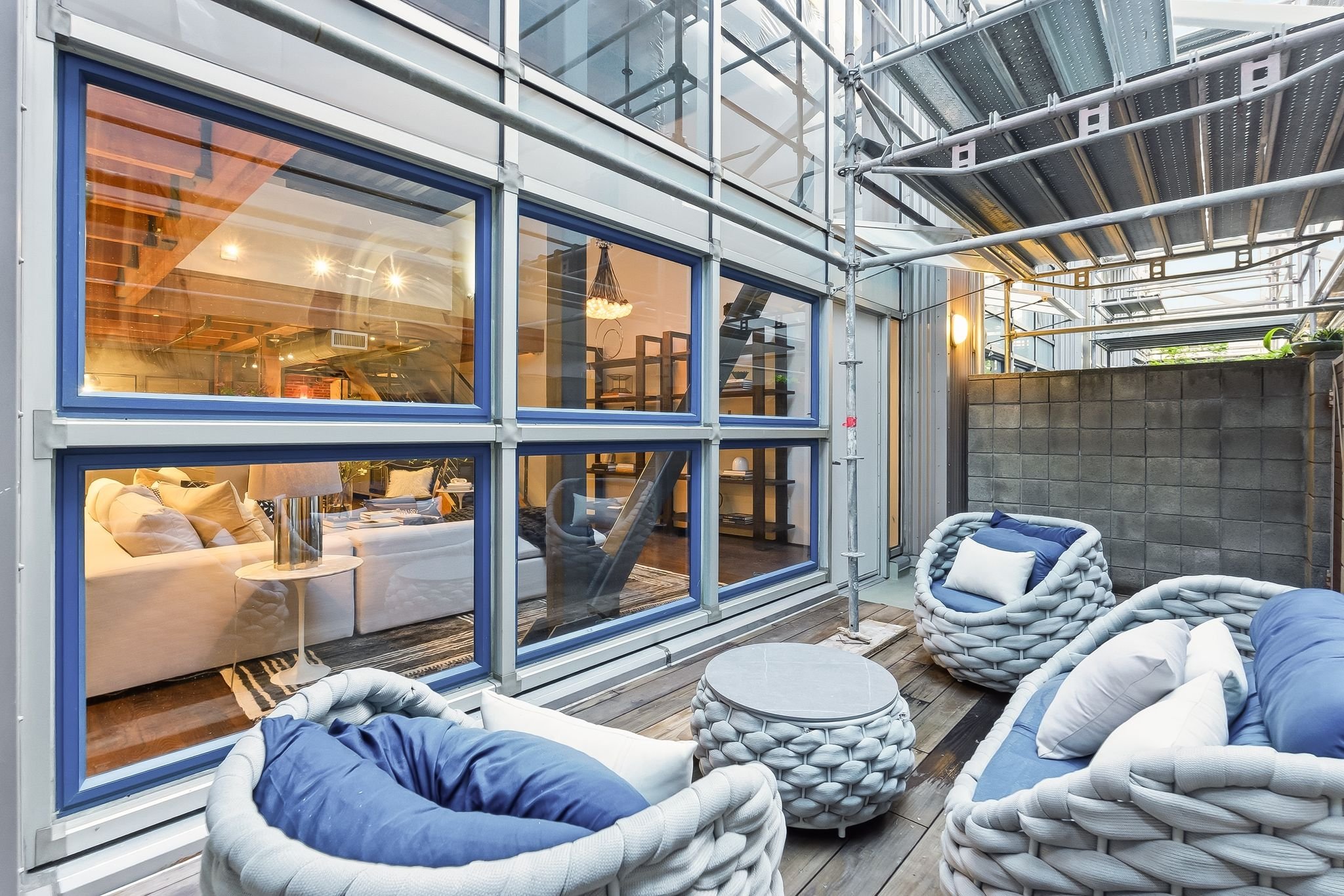 Exterior balcony with modern glass walls, outdoor furniture including cushioned chairs and small tables, and scaffolding for construction or maintenance.