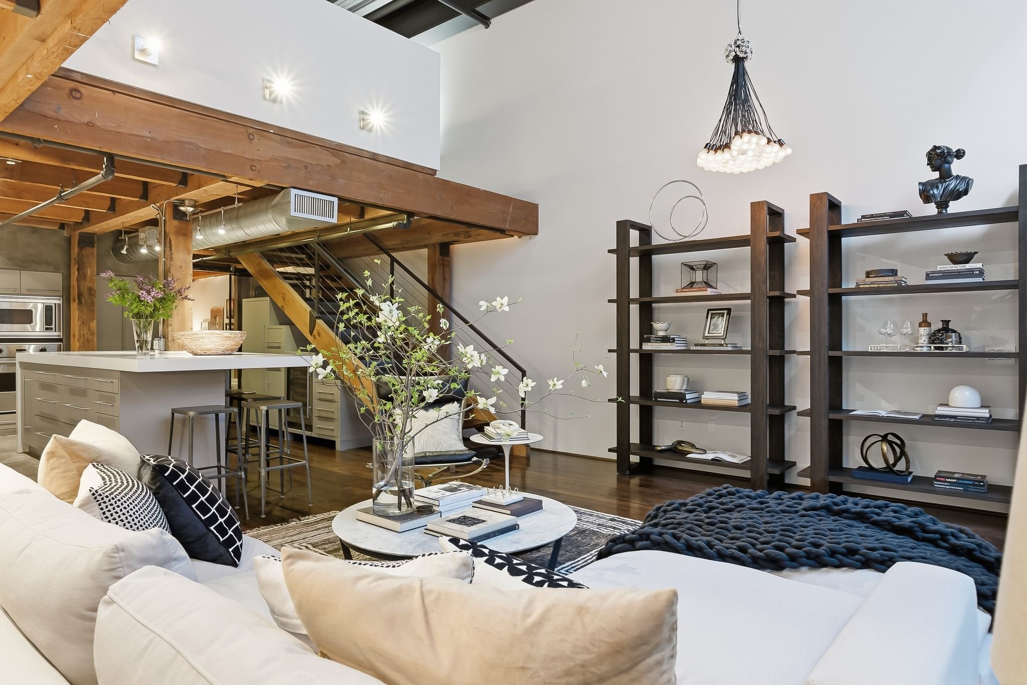 Open-concept living space with a white sectional sofa accented by black and white pillows, a round coffee table with books, a dark textured ottoman, and a large modern chandelier. A bookshelf against the wall displays books and decorative objects. Th