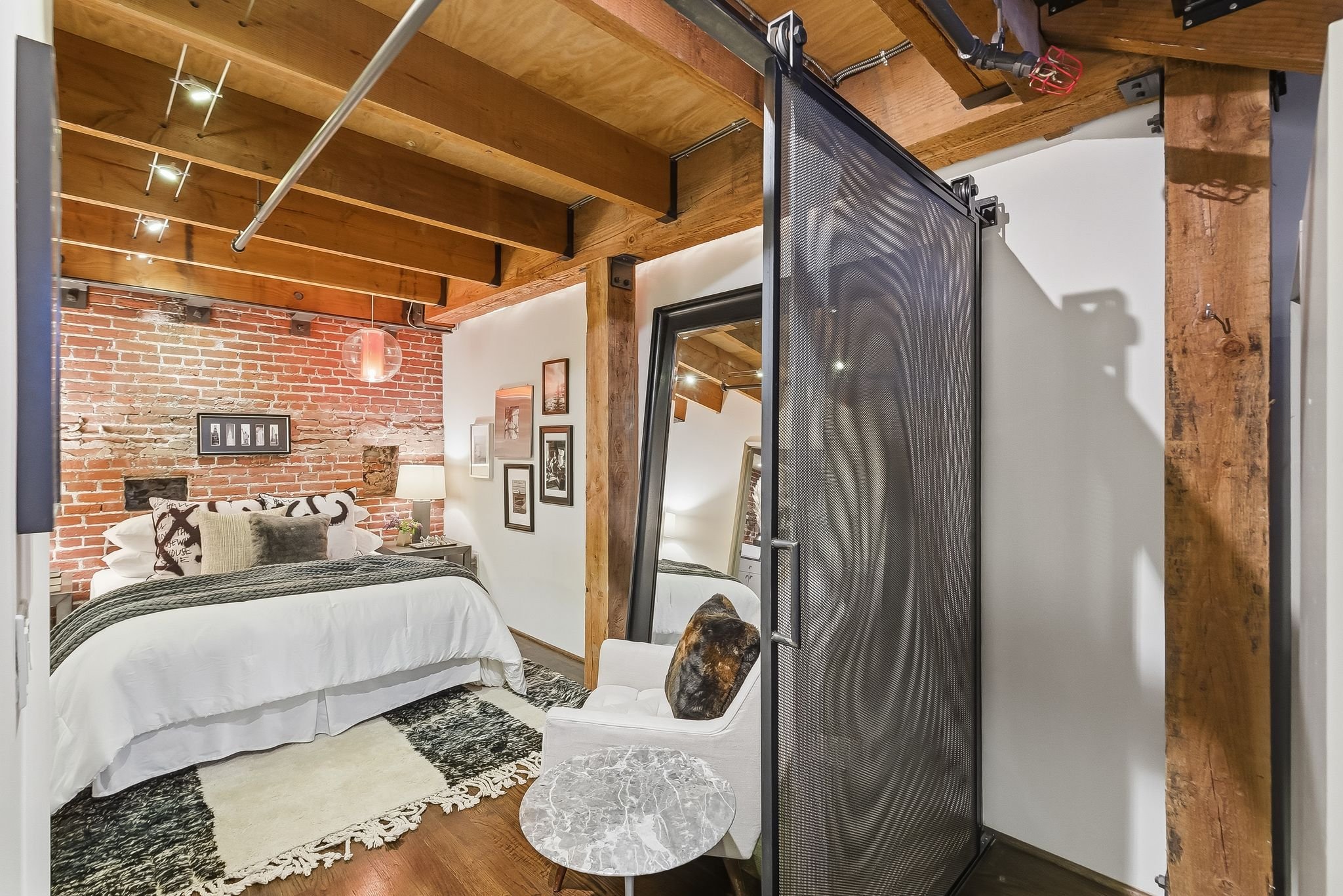 A cozy bedroom with a brick accent wall, a white bed with decorative pillows and a black and white rug, framed photos on the wall, wooden ceiling beams, and a glass table next to a white chair with a dog resting on it. There is a black metal room div