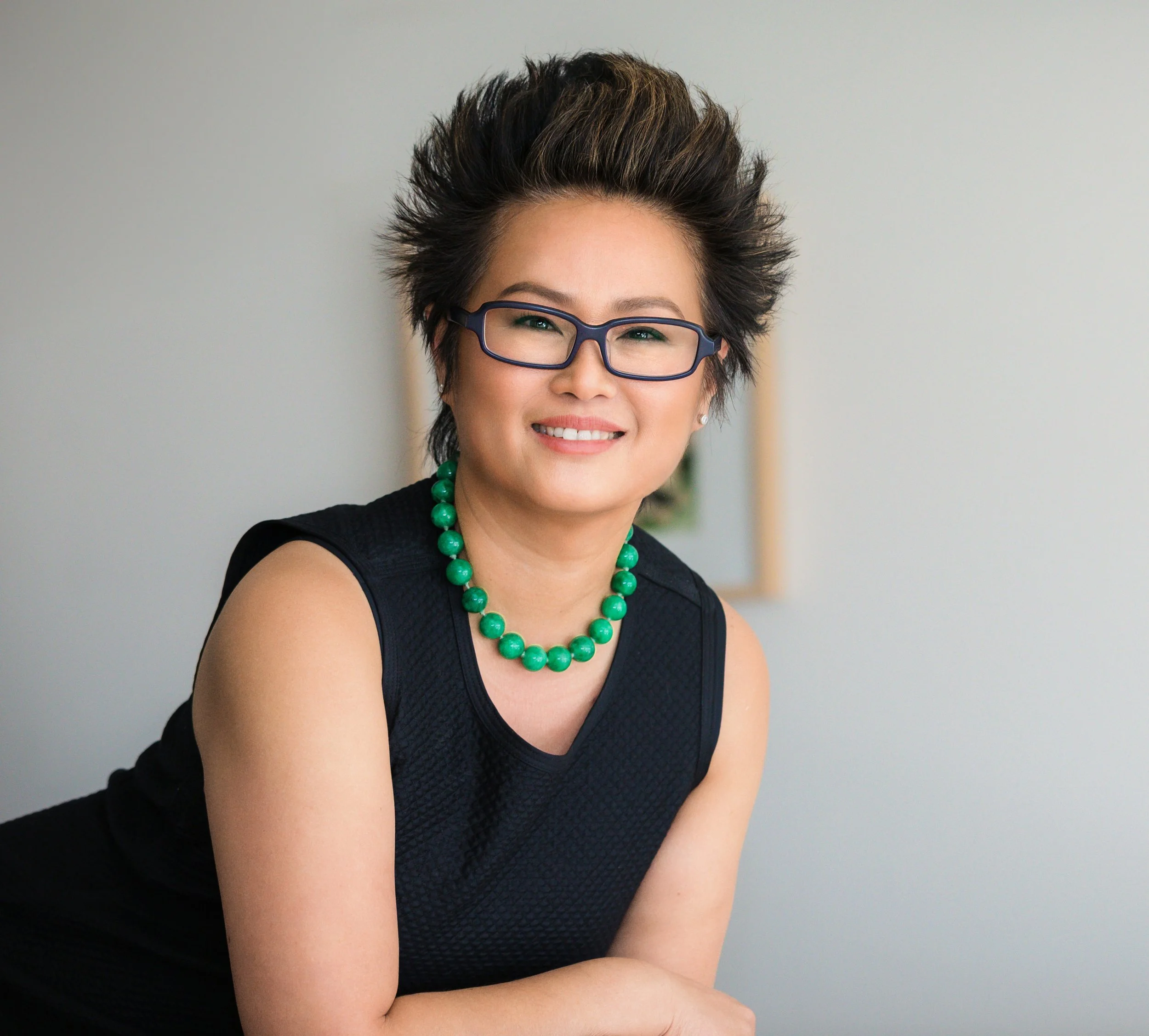 A woman smiling, wearing glasses, a sleeveless black top, and a green beaded necklace, with short spiked hair."}