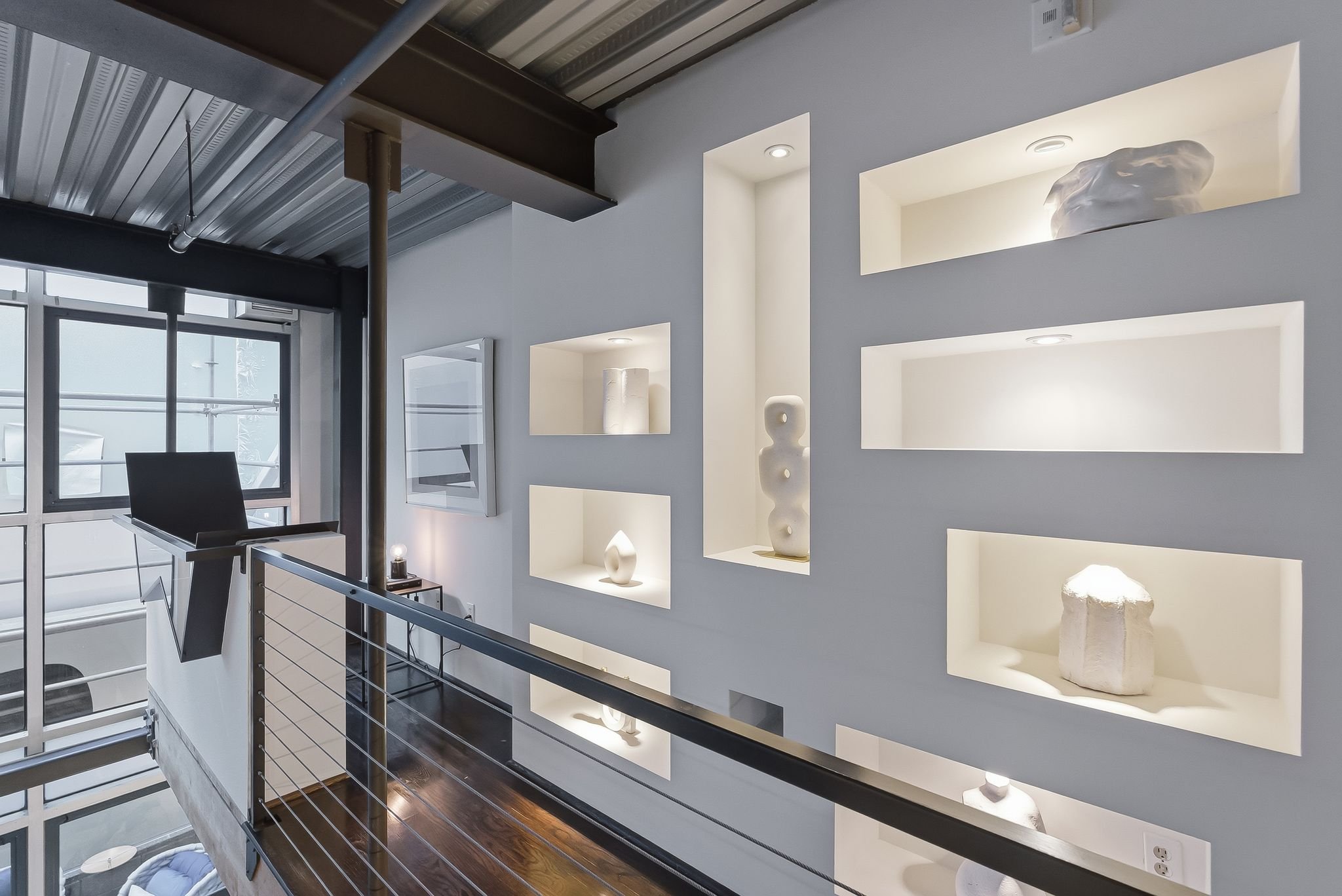 Interior view of a modern building with a metal ceiling, large window, and decorative white display shelves built into the wall, showcasing various white sculptures and vases.