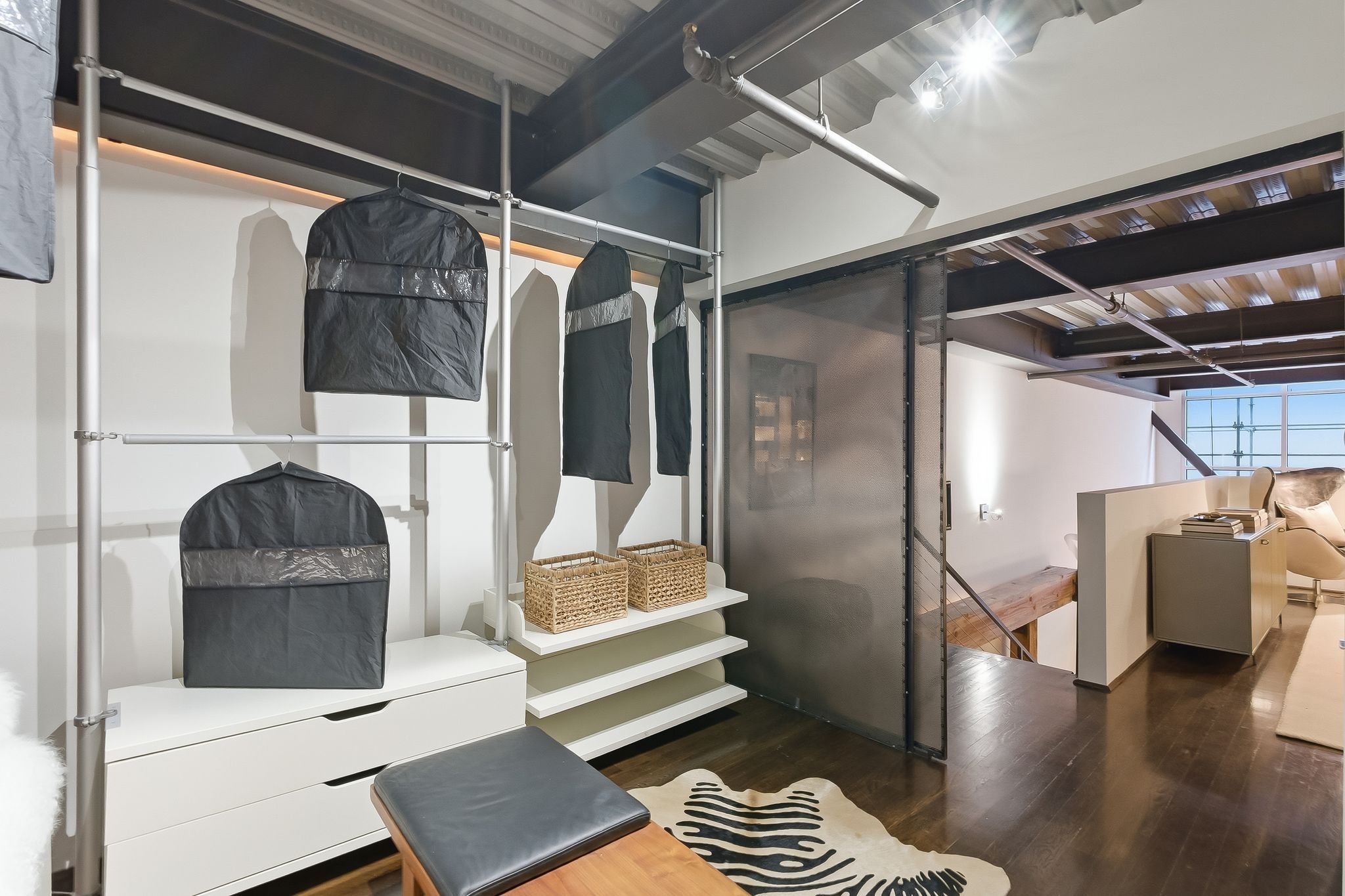 Interior view of a modern living space showing a clothing rack with black bags and hanging clothes, a white shelf with wicker baskets, a black cushioned stool, and a staircase leading to an upper level with visible railing. The room has wood floors a