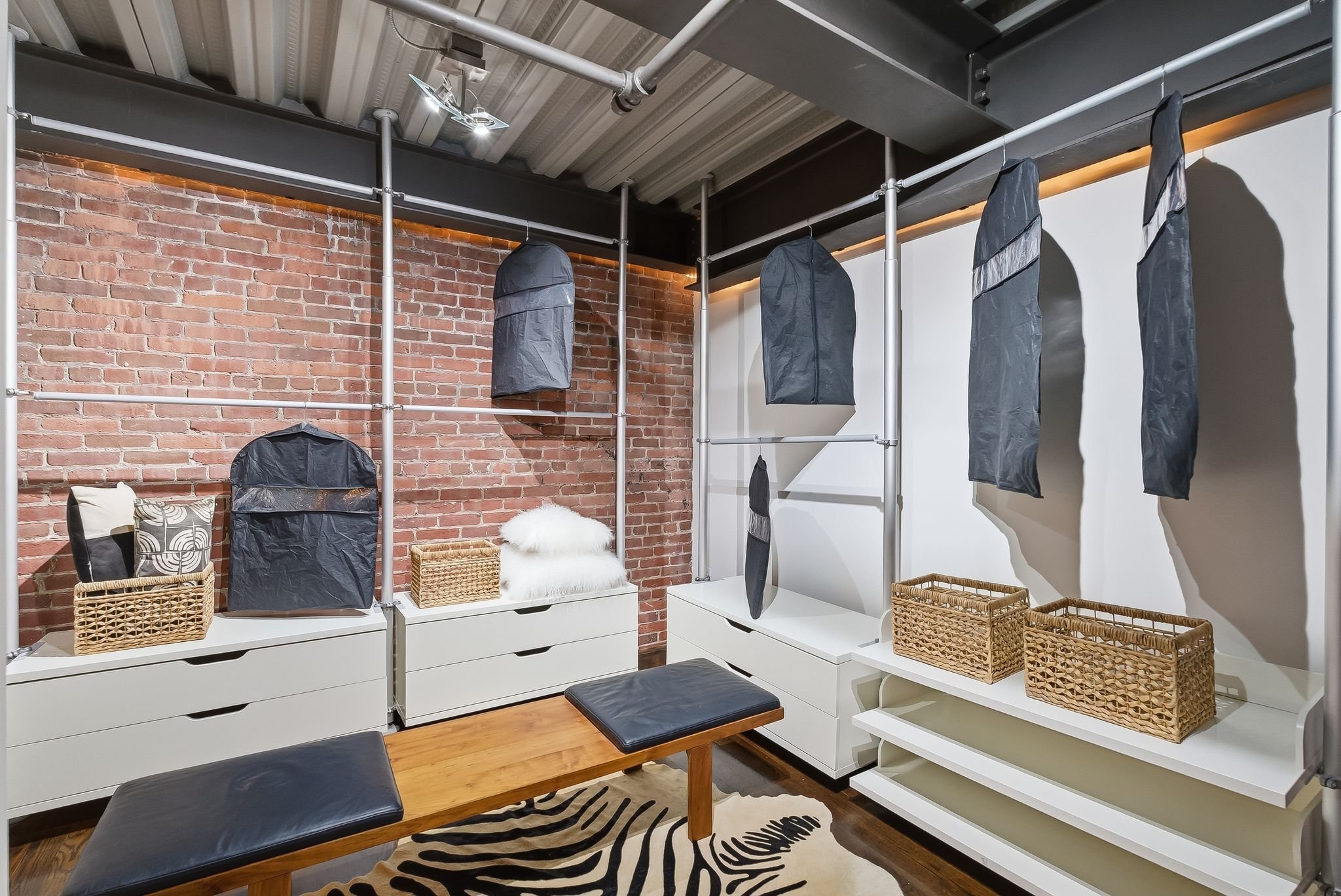 Closet with hanging clothing covered in garment bags, wicker baskets, and white storage drawers against a brick wall and a white wall, with a wooden bench and a zebra-patterned rug on the floor.