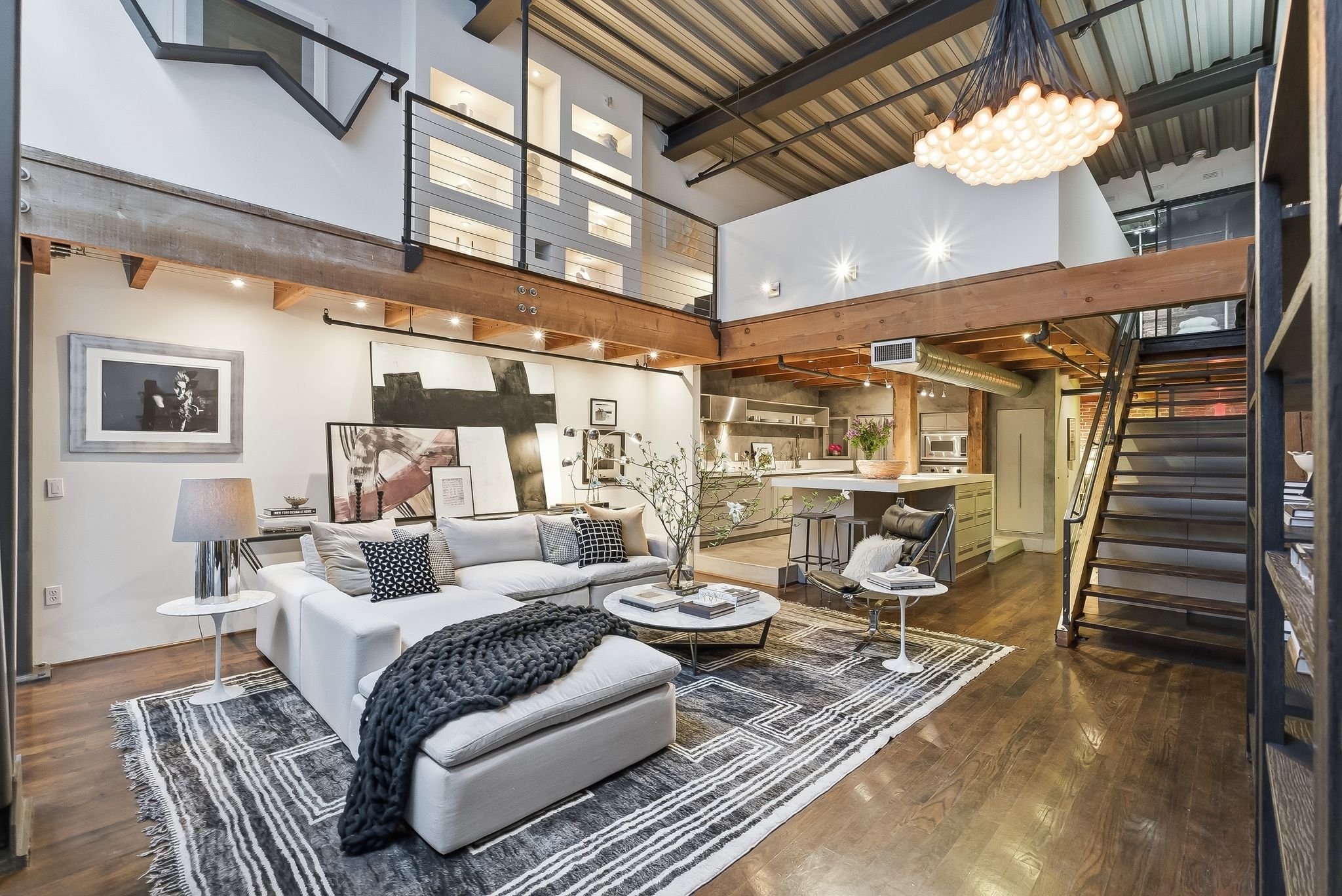 Open-concept loft with modern industrial decor, featuring a white sectional sofa with black and white throw pillows, a black chunky knit blanket, a striped rug, a round coffee table, and a kitchen with a large island and stainless steel appliances. E