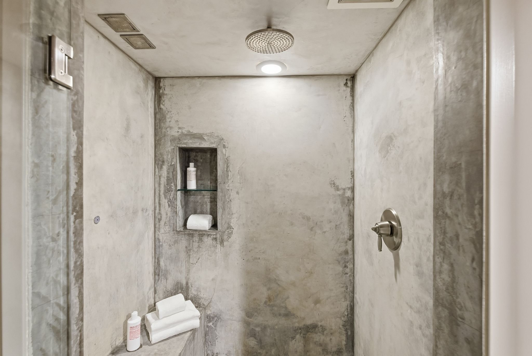Empty shower with concrete walls, built-in shelf holding shampoo and towels, showerhead, and a bottle of body wash.