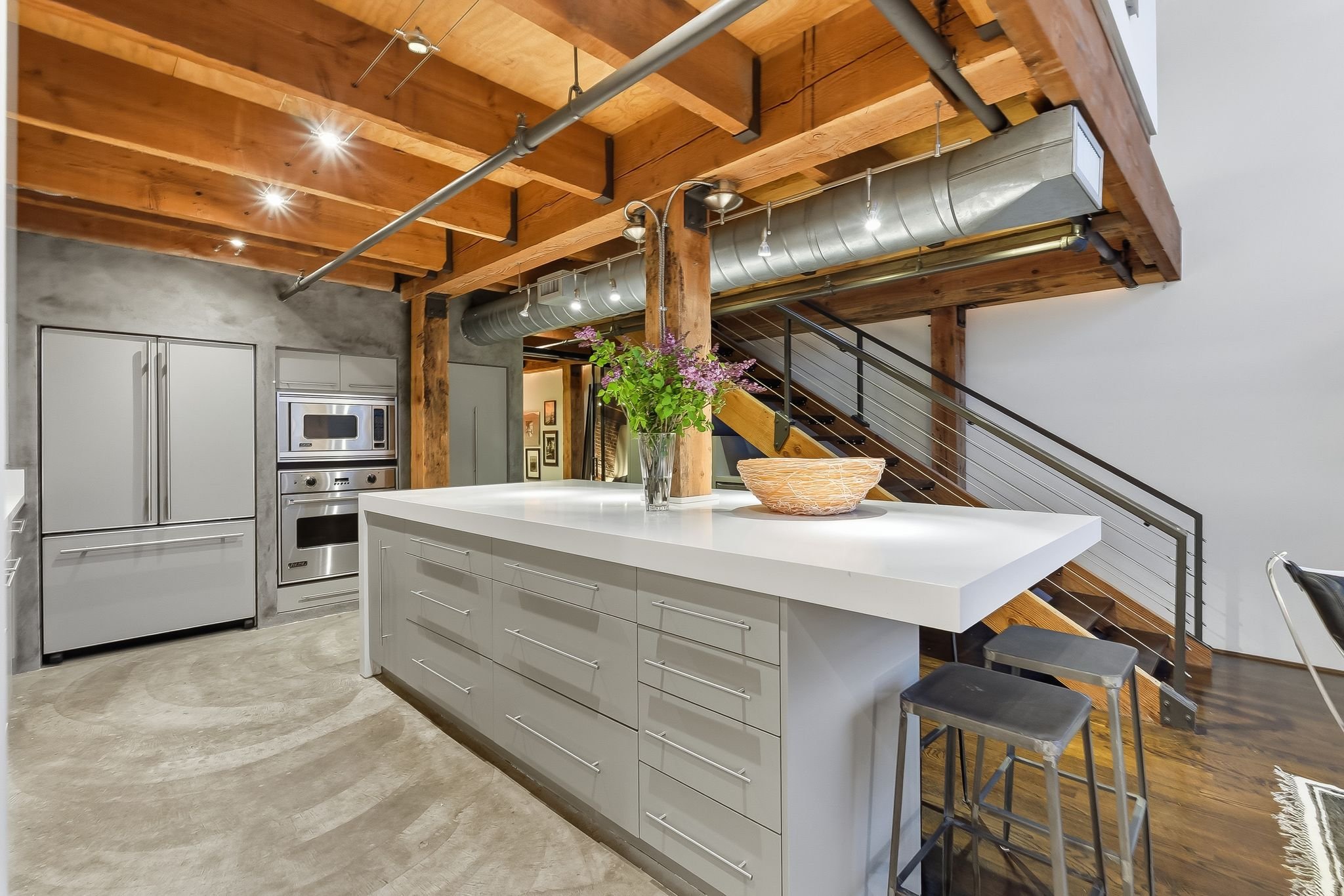 Modern kitchen with a white island, gray appliances, wooden beams, stairway, and industrial-style ceiling pipes.