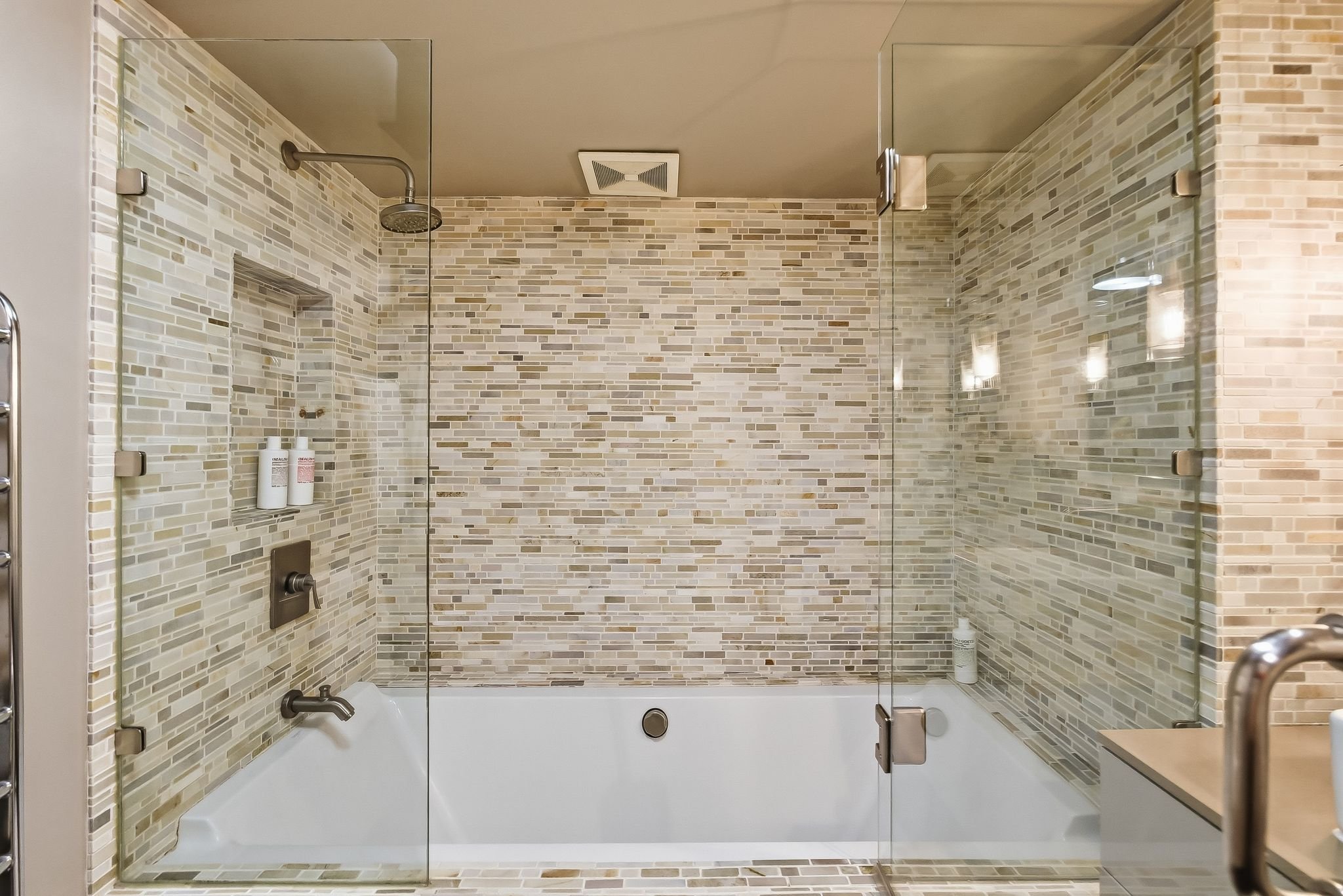 A modern walk-in shower with glass doors, beige and brown mosaic tile walls, a showerhead, and a white bathtub with a wall-mounted faucet.