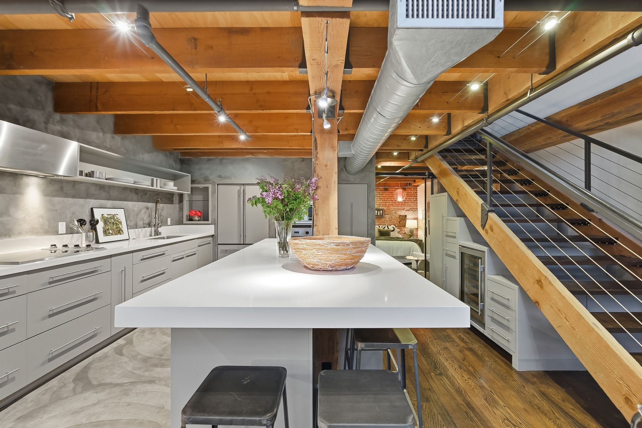 Modern kitchen with white cabinets, a large white island, a flower vase, exposed wooden beams on the ceiling, ductwork, a staircase with black railings, and a brick wall in the background.