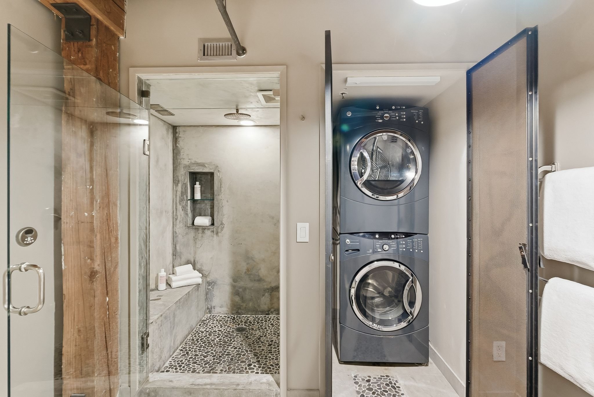 A laundry room with a front-loading washer and dryer stacked beside a gray wall. There is a metal shelf and a black mesh laundry hamper. The room has a small pebble-patterned floor and a doorway leading to a bathroom with a shower and shelves with to