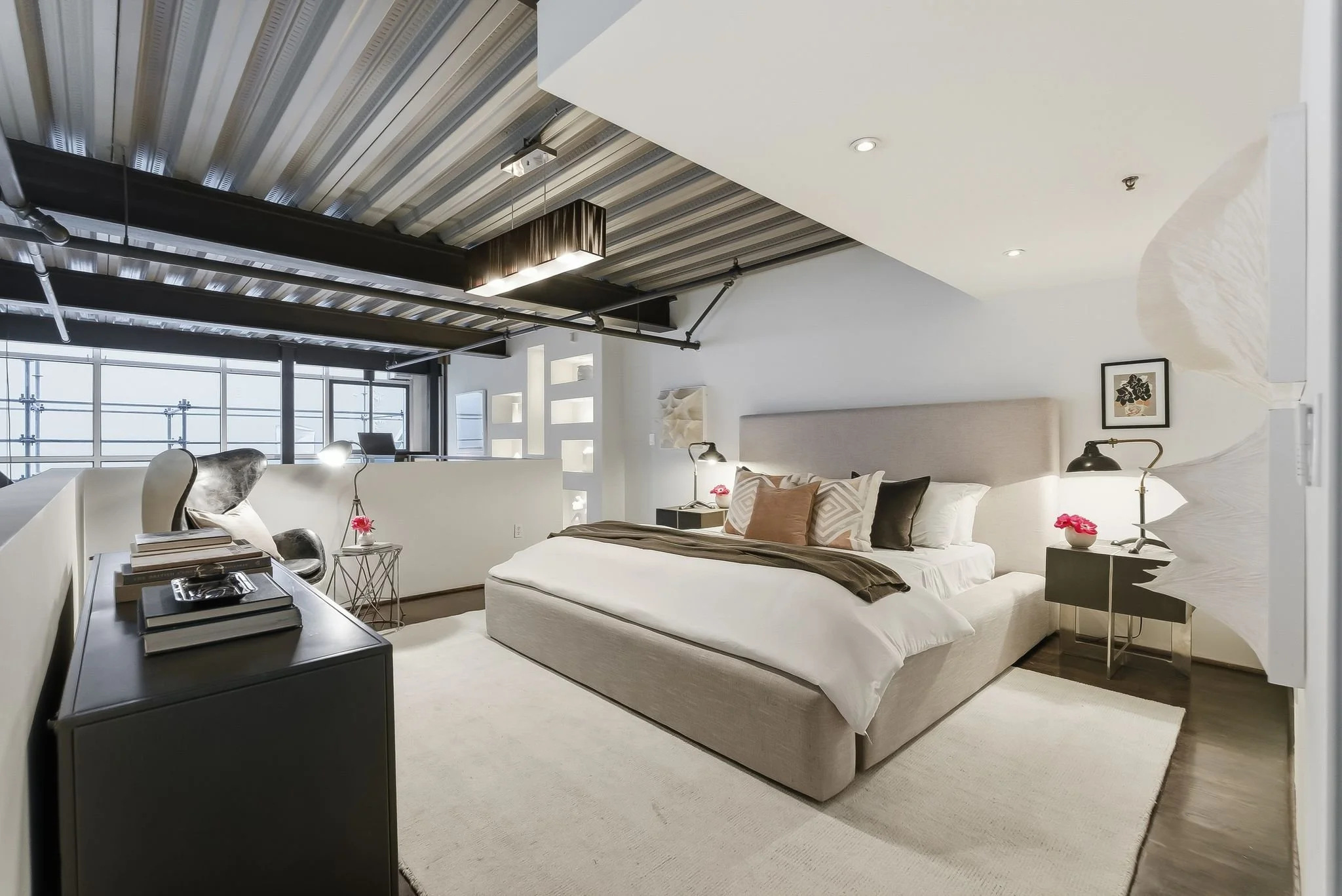 A modern, minimalistic bedroom with a large bed, pillows, and bedside lamps, featuring an industrial ceiling with exposed pipes and ductwork, and large windows letting in natural light.