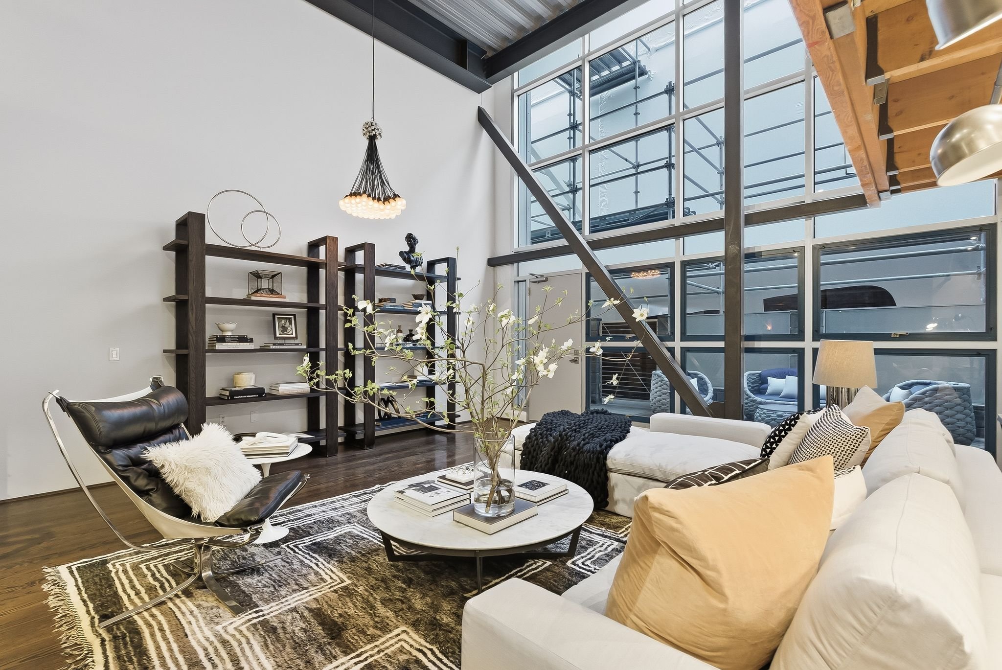 Modern living room with large floor-to-ceiling windows, featuring a white sofa with pillows, a round coffee table with a flower arrangement, a black lounge chair with a faux fur pillow, a wooden bookshelf, a unique chandelier, and a patterned rug.