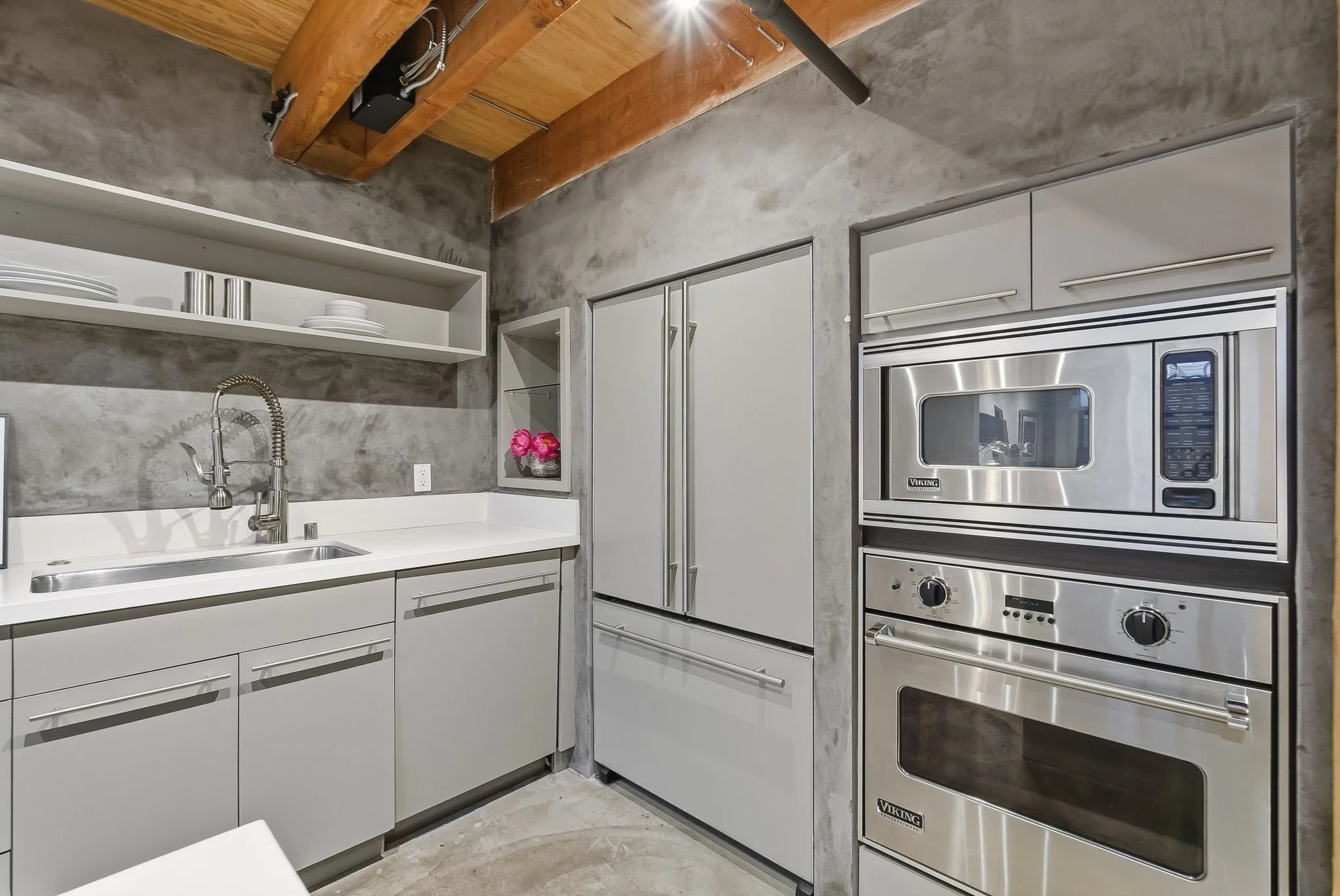 Modern kitchen with concrete wall finish, white cabinets, open shelf with dishes, stainless steel oven and microwave, and a sink with a modern faucet.