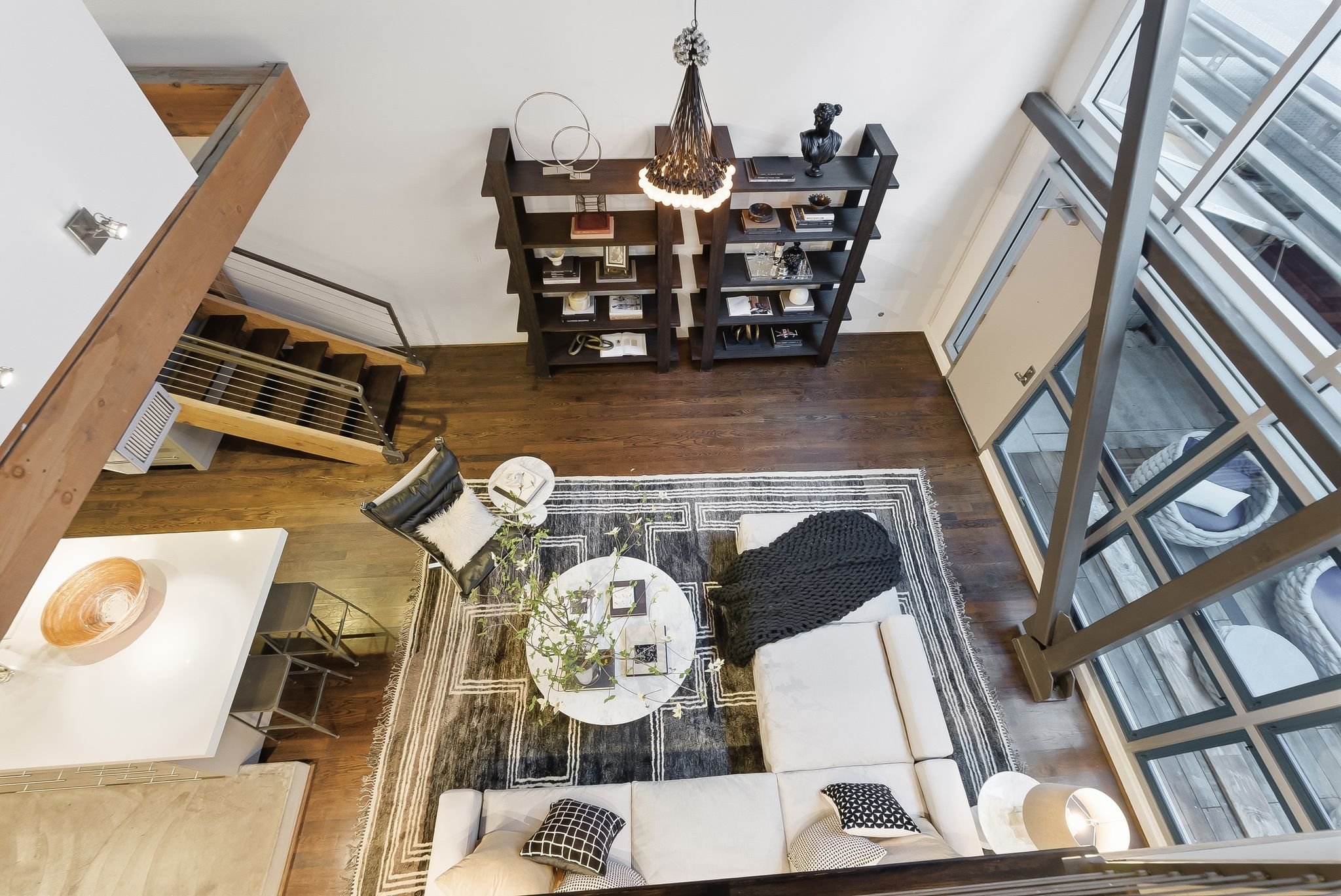 Living room with dark wood bookshelves, a black pendant light, glass and decorative items, a white sofa with black and white pillows, a black and white area rug, a black leather armchair with a white pillow, and large windows letting in natural light