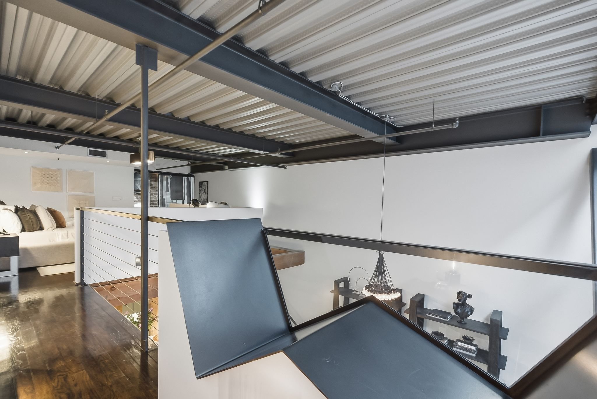 Modern interior with a ceiling made of corrugated metal panels, visible steel beams, and hanging piping; a lounge area with a beige sofa and decorative pillows, and a dining area with a round chandelier and a wooden table.