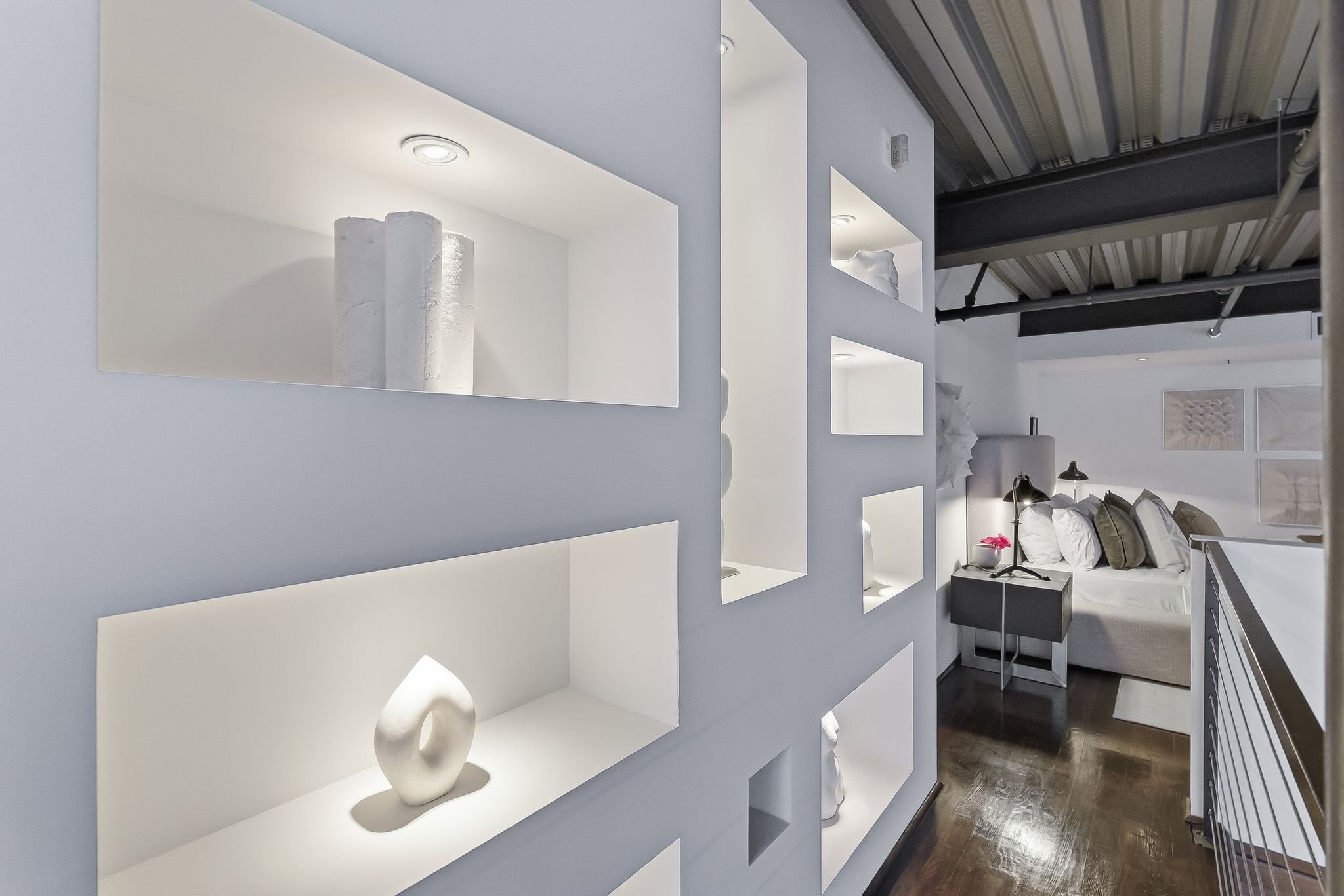 A modern, minimalist bedroom with a white built-in shelf featuring decorative white vases and lights. The room has a bed with white bedding, pillows, and a nightstand with a pink flower in a white pot. Exposed industrial-style ceiling with pipes and 
