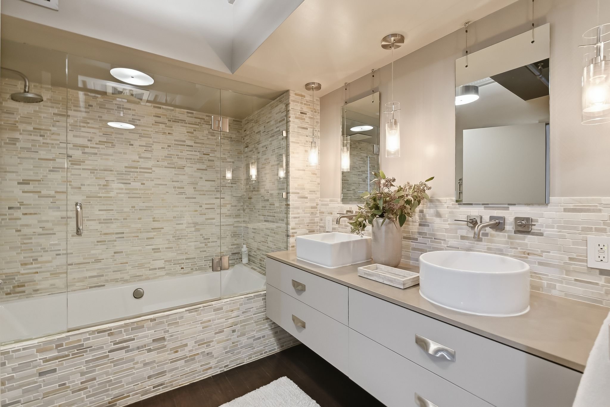 Modern bathroom with glass-enclosed shower, mosaic tile walls, white vanity with vessel sinks, and decorative hanging lights, with a mirror and plant on the countertop.