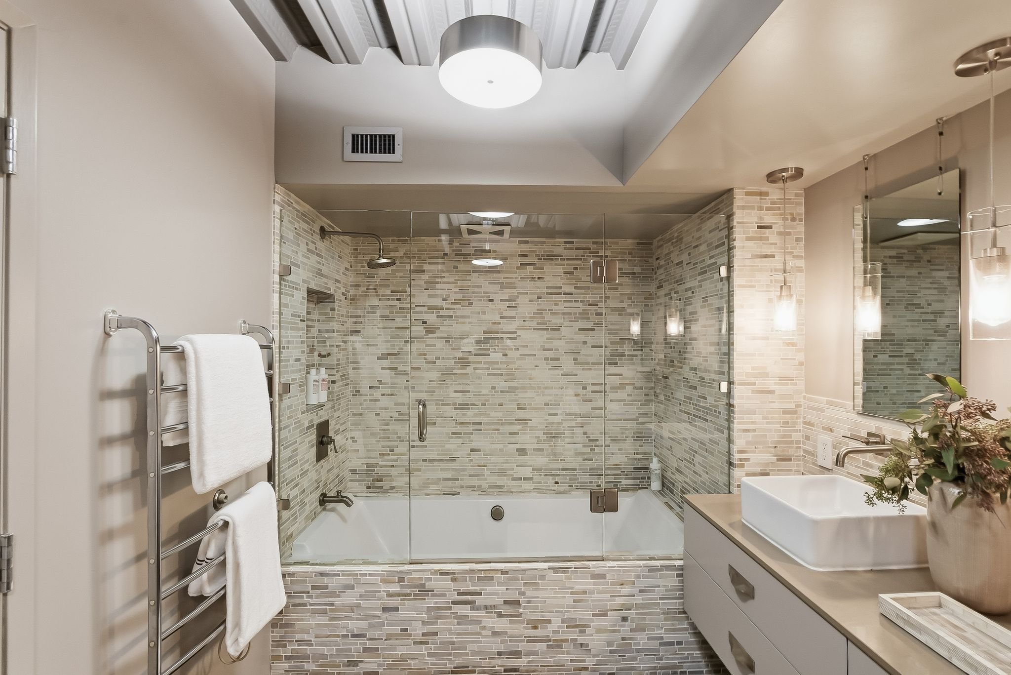 Modern bathroom with a glass shower enclosure, tiled walls, a white bathtub, a wall-mounted sink with a potted plant, and a heated towel rack.