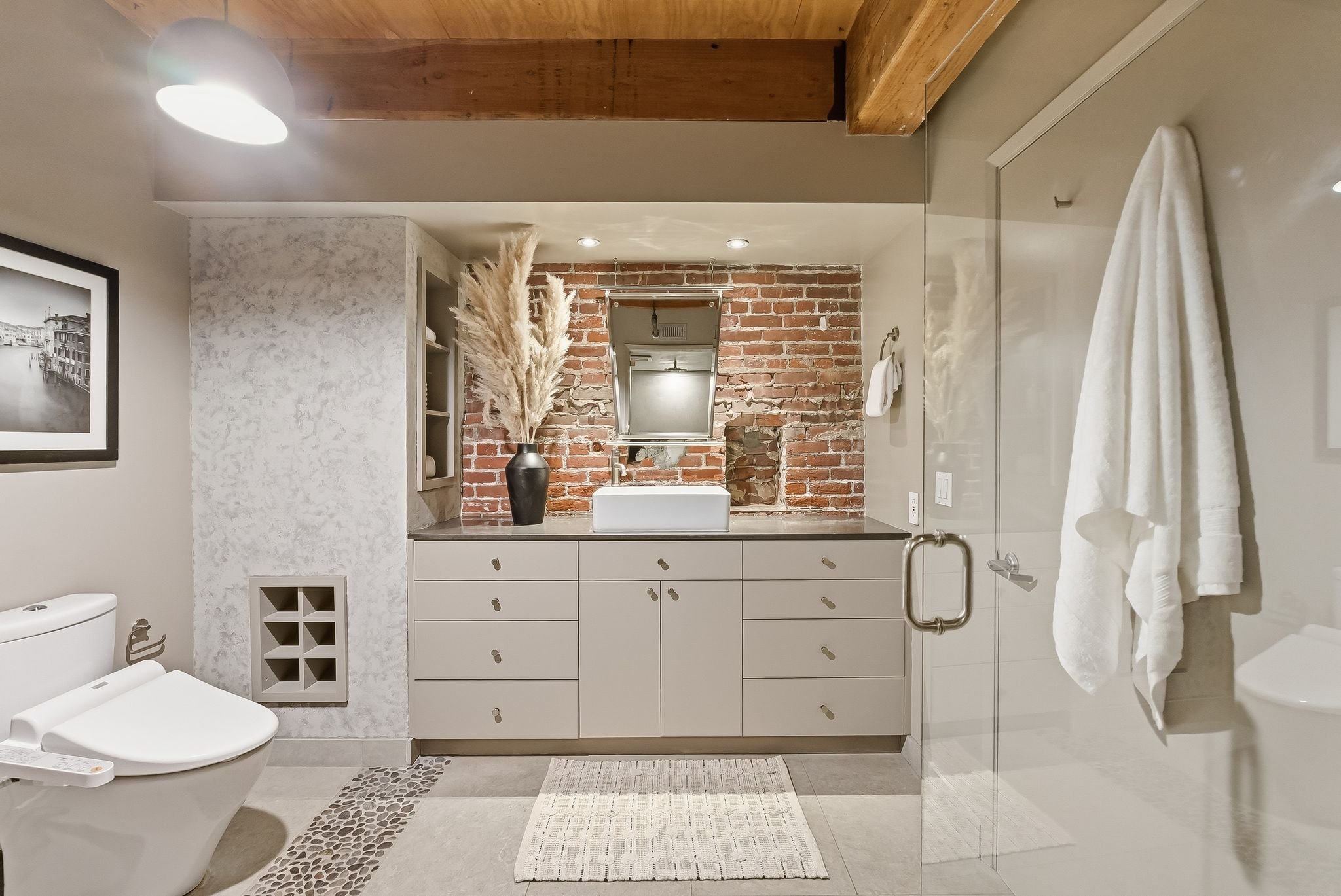 Modern bathroom with a brick wall behind a white sink and cabinetry, decorative dried pampas grass in a vase, a mirror, and a toilet, with neutral-colored towels and a glass shower door.