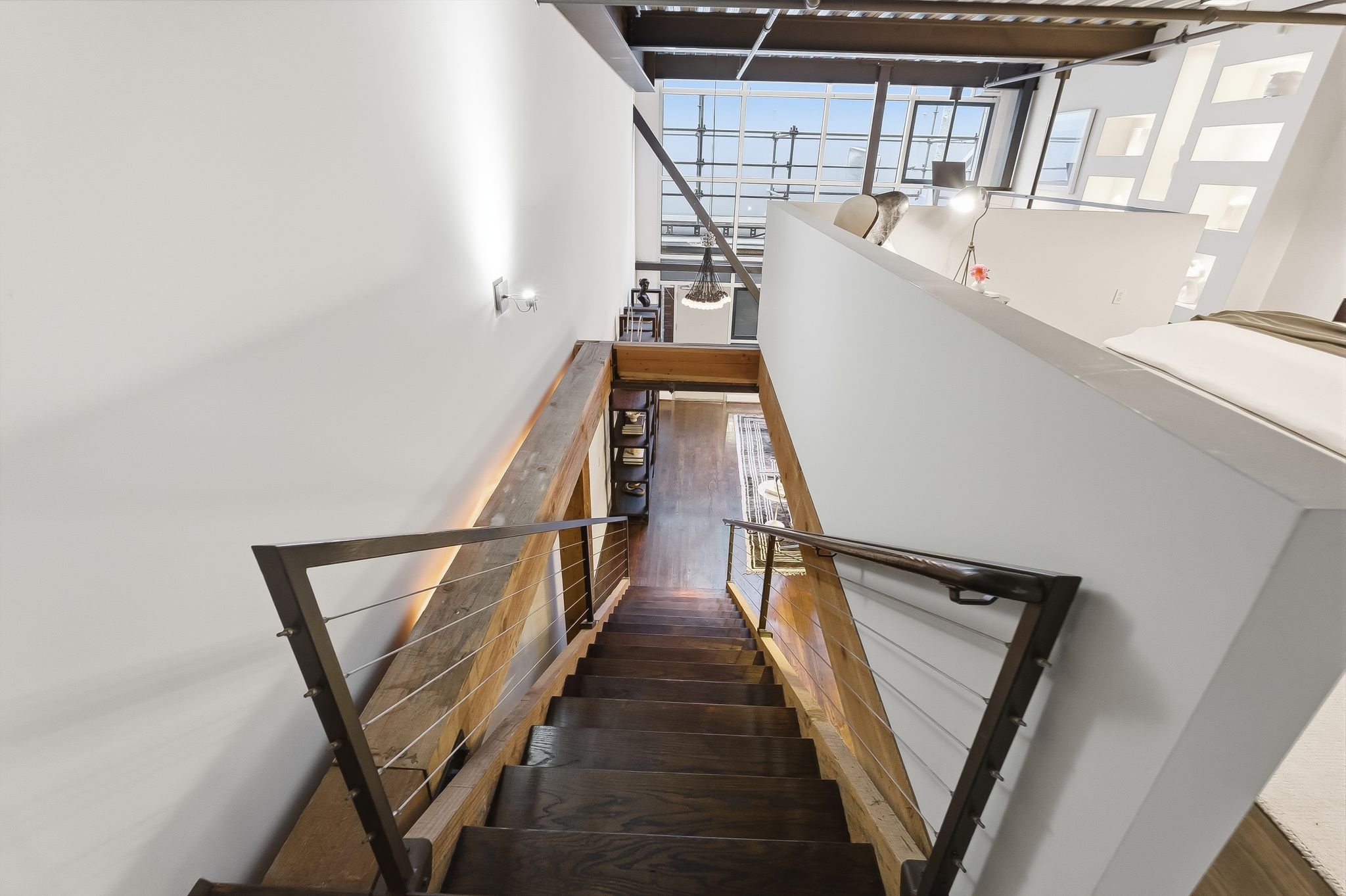 View looking down a staircase in a modern, bright interior with white walls, wooden stairs, and metal railings, overlooking a living space with large windows and furniture.