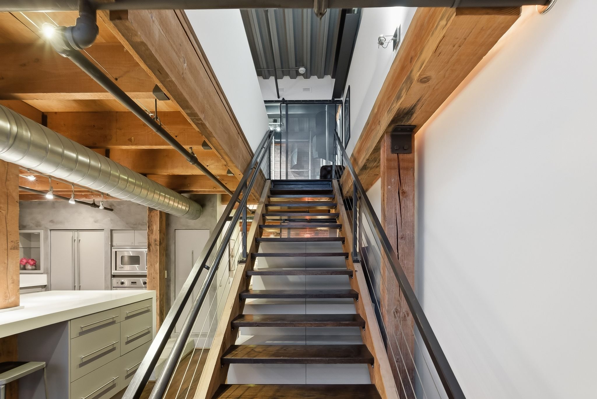 Interior view of a modern staircase with metal railings, wooden steps, and walkways leading to an upper level with glass door, in an industrial style space with exposed ductwork and wood accents.