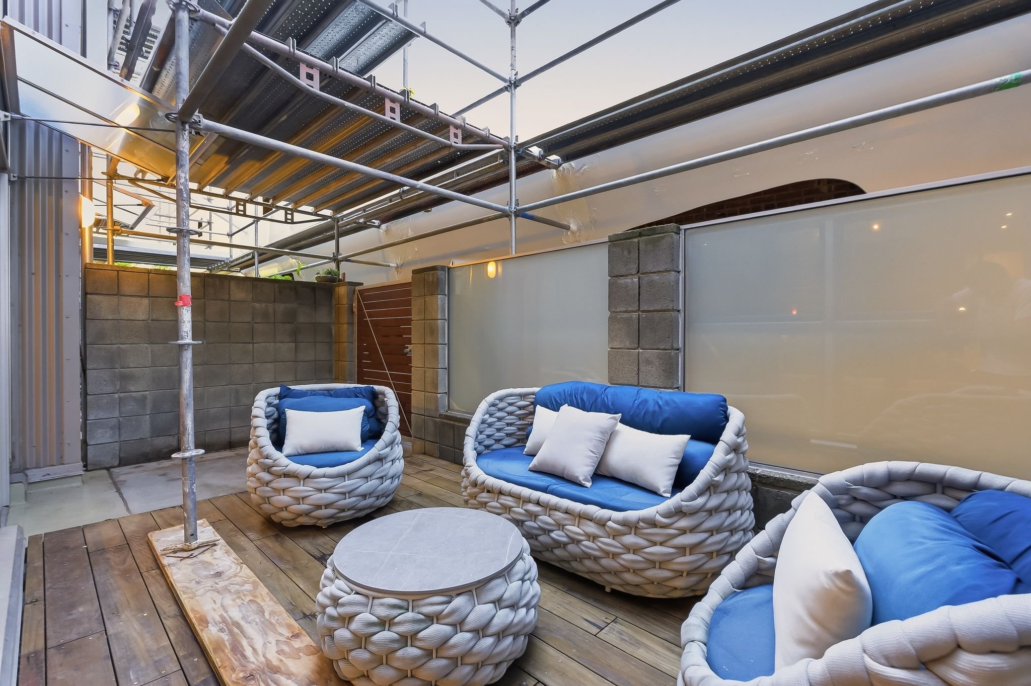 An outdoor seating area with wicker chairs with blue cushions and white pillows, a round white table, a wooden deck, and scaffolding above.