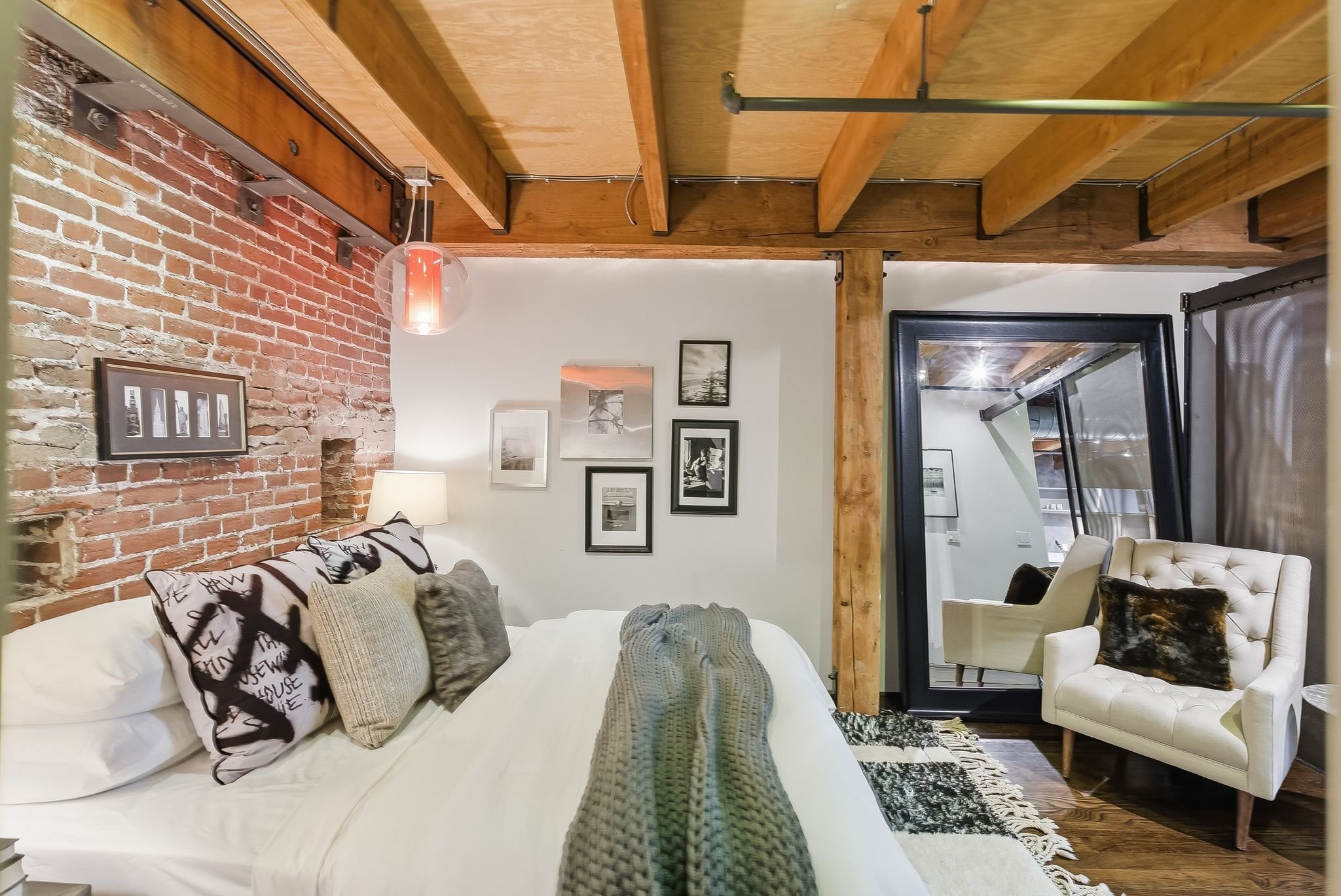 A cozy living room with a white sofa decorated with patterned cushions, a tufted white armchair, a black and white rug, and framed photos on the wall. Exposed brick wall and wooden beams add rustic charm.