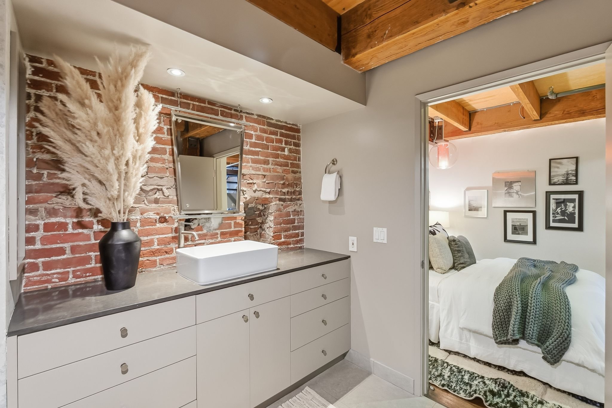 Bathroom with a white sink on a countertop, a black vase with pampas grass, a mirror on an exposed brick wall, and a bedroom with a white bed and framed pictures on the wall visible through an open doorway.