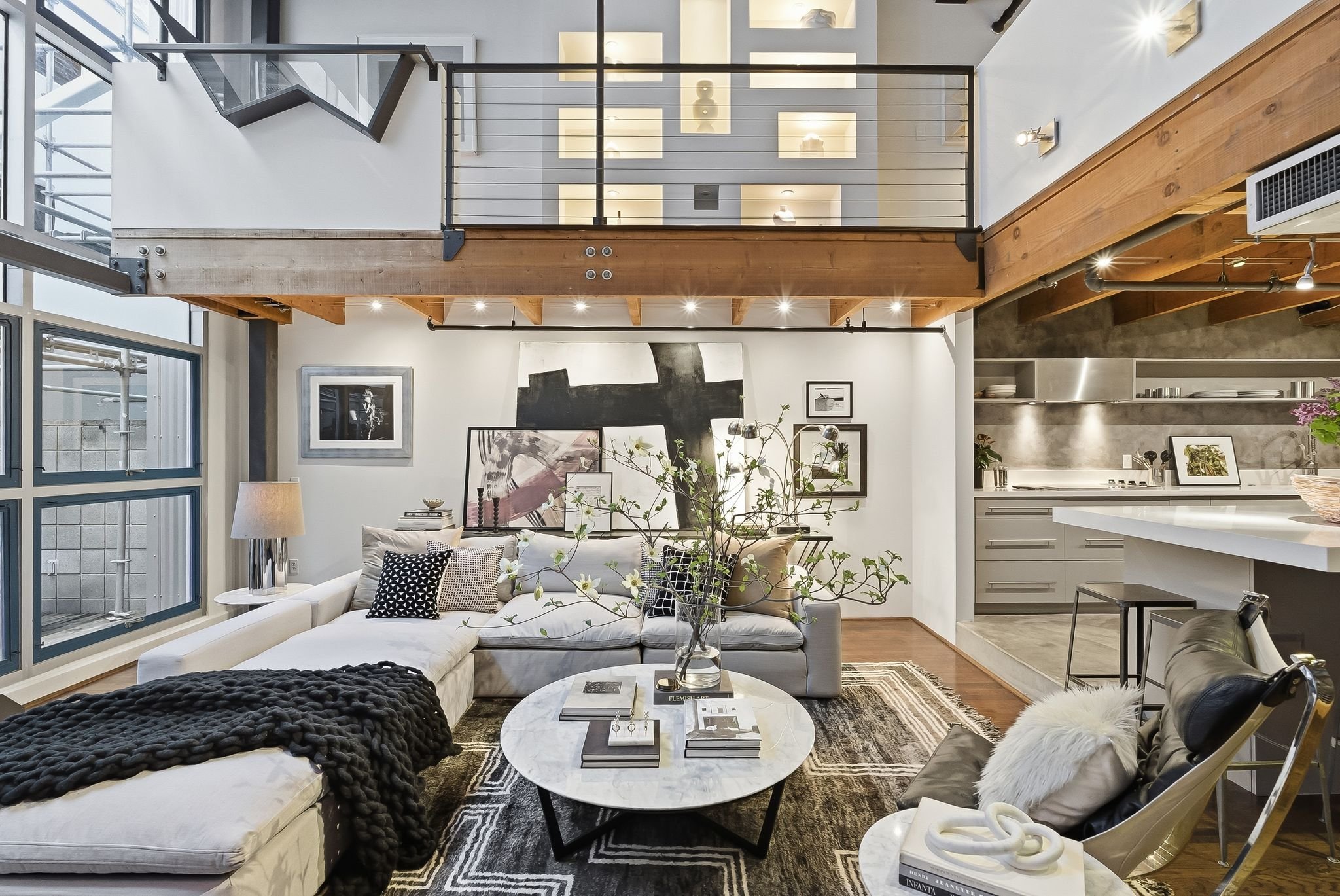 Interior view of a modern living room with white sofa, black and white pillows, artwork on the wall, a marble coffee table with books and decorative items, and a large window with glass doors leading outside. The room has wooden beams and a kitchen a