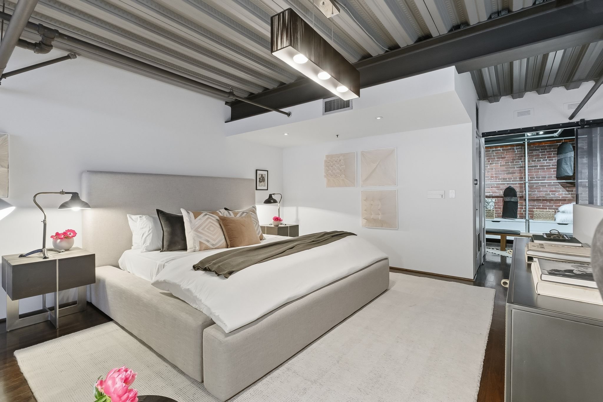 Modern bedroom with a large bed, beige upholstered headboard, black and beige accent pillows, and a beige blanket. Two nightstands with lamps and pink flowers flank the bed. White walls feature framed artwork, and there is a door leading to a brick p
