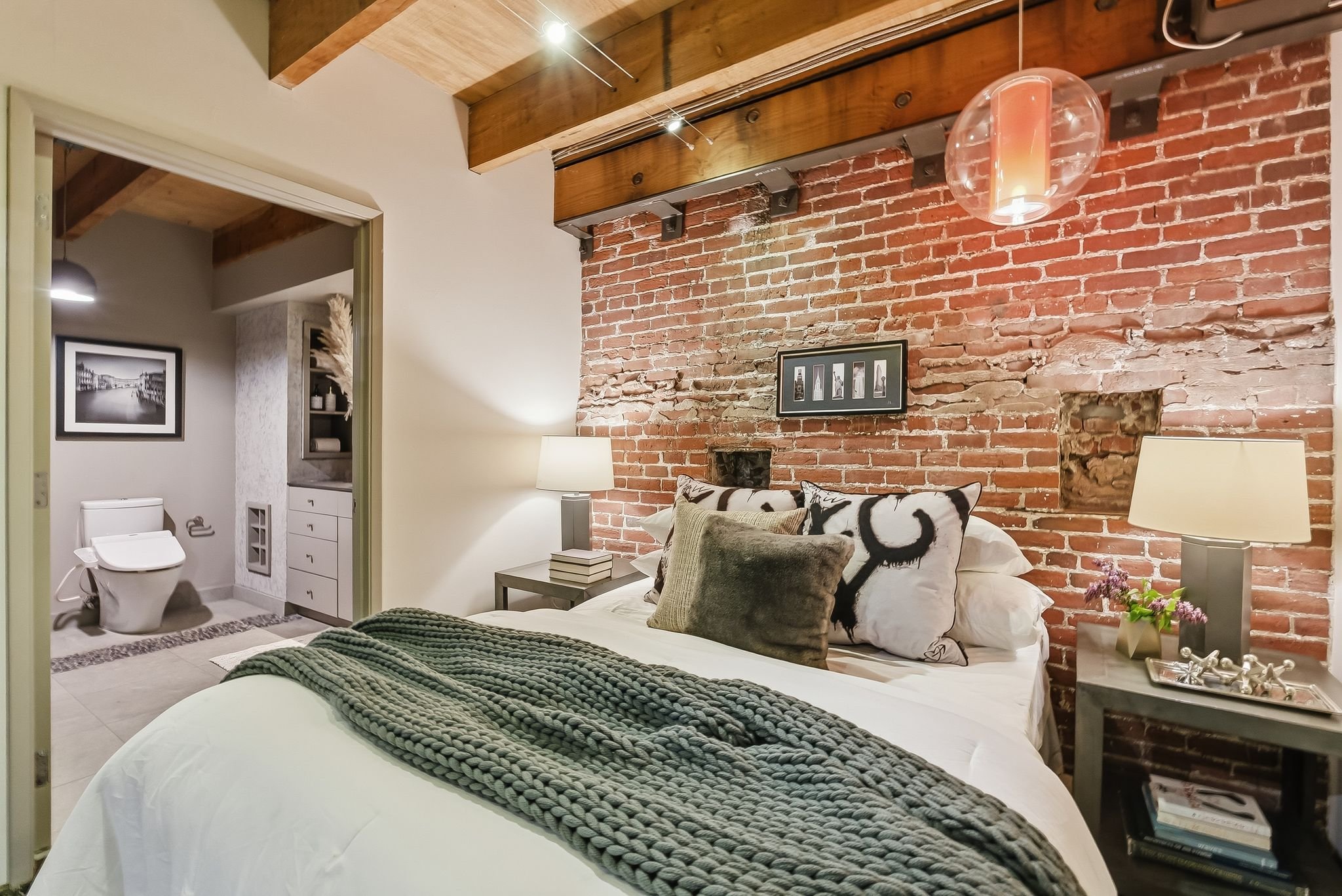 A bedroom with an exposed red brick wall, a bed with a textured throw blanket, and decorative pillows. There are two nightstands with lamps, a framed picture above the bed, and a small plant on one nightstand. A pathway leads to a bathroom visible th