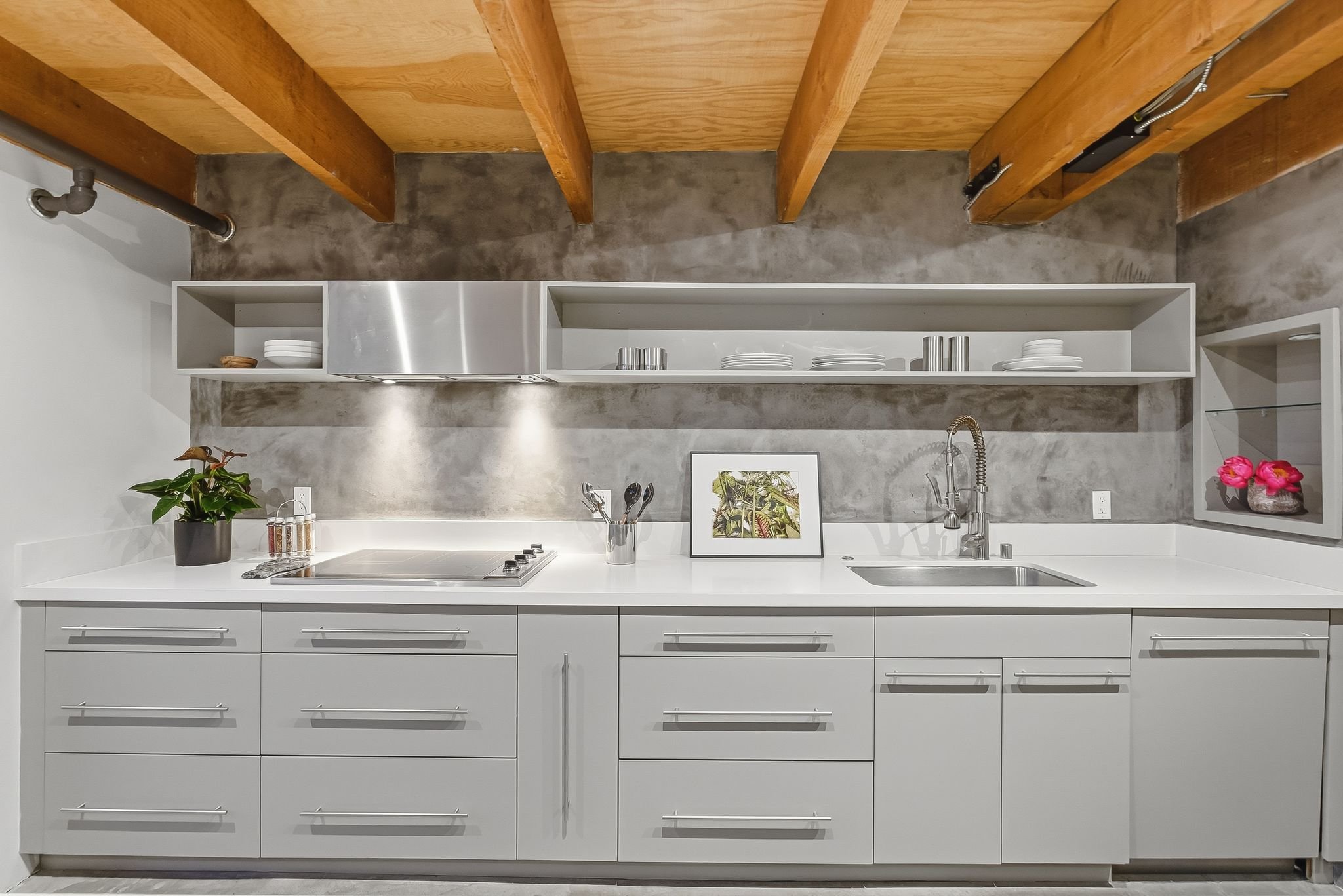Modern kitchen with white cabinetry, a concrete backsplash, a sink, a framed picture, a potted plant, and open shelves with plates and utensils. Exposed wooden ceiling beams and a gray floor.
