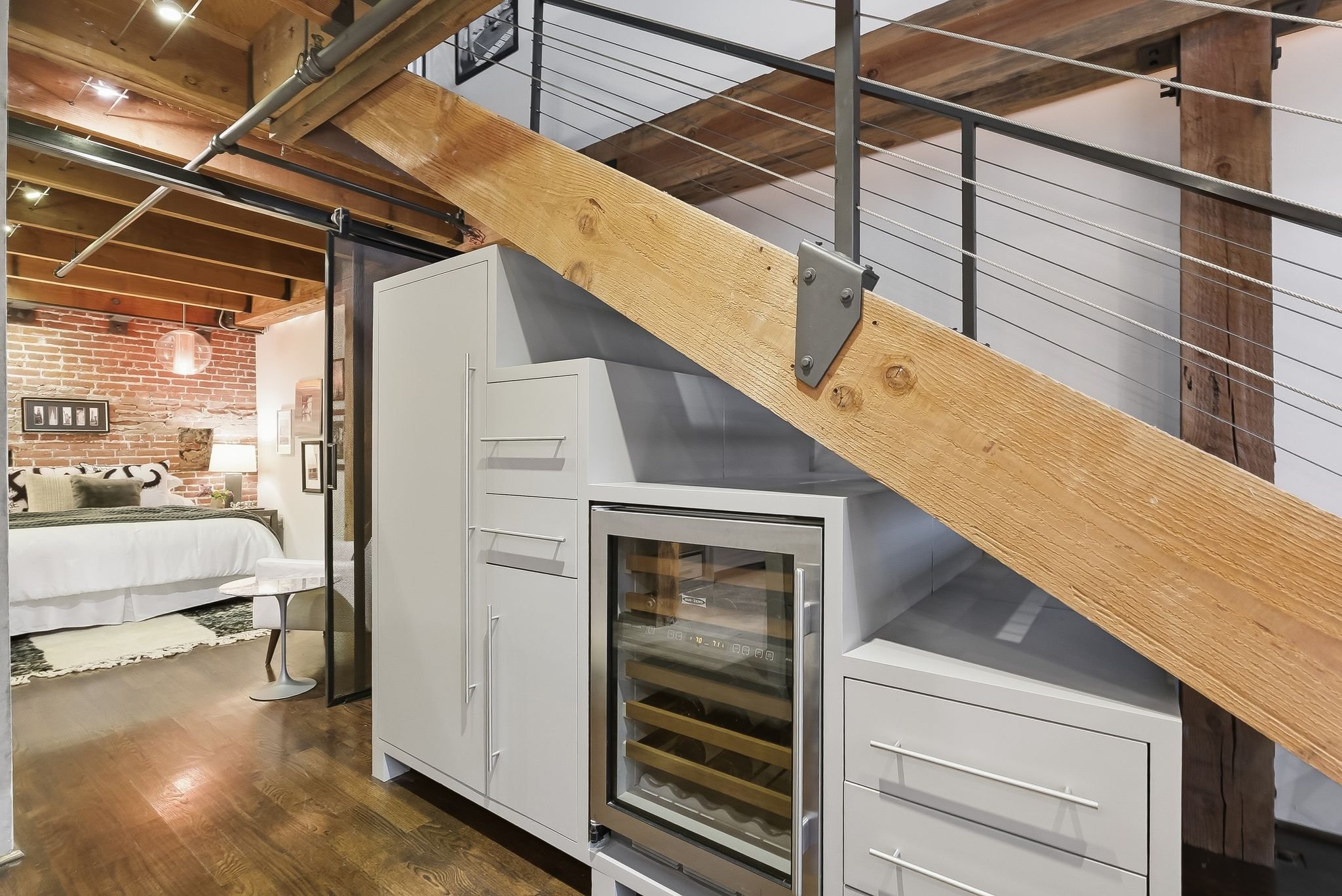 Interior view of a modern loft with exposed brick wall, a white bed with black and white pillows, a small round side table, and a kitchen area with white cabinets and a wine cooler, featuring a wooden staircase with metal railing and cables.
