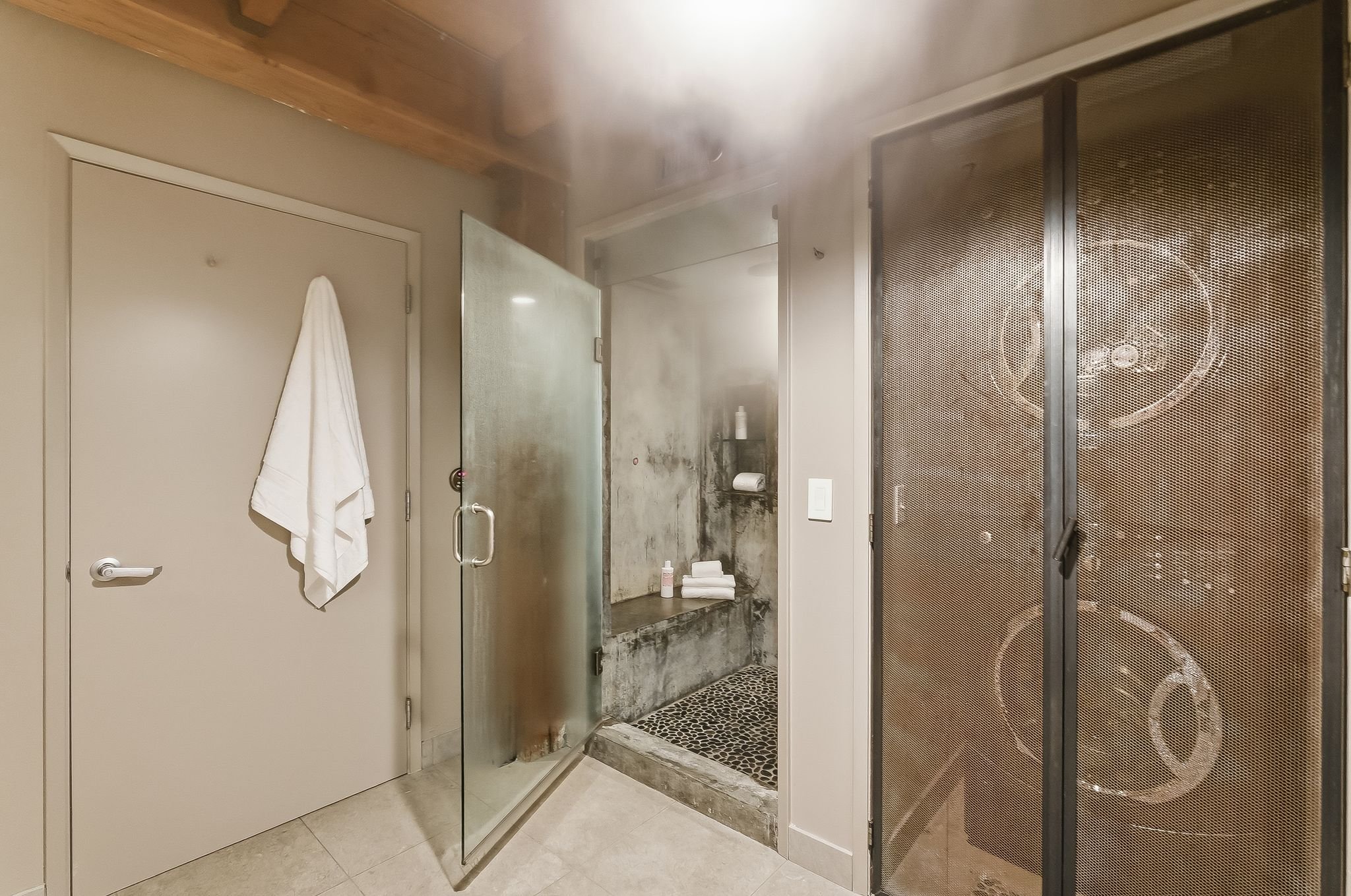 Inside a bathroom, showing a closed door with a towel hanging on the handle, a frosted glass shower door, a shower area with a bottle and folded towels, and a metal cabinet with patterned doors.