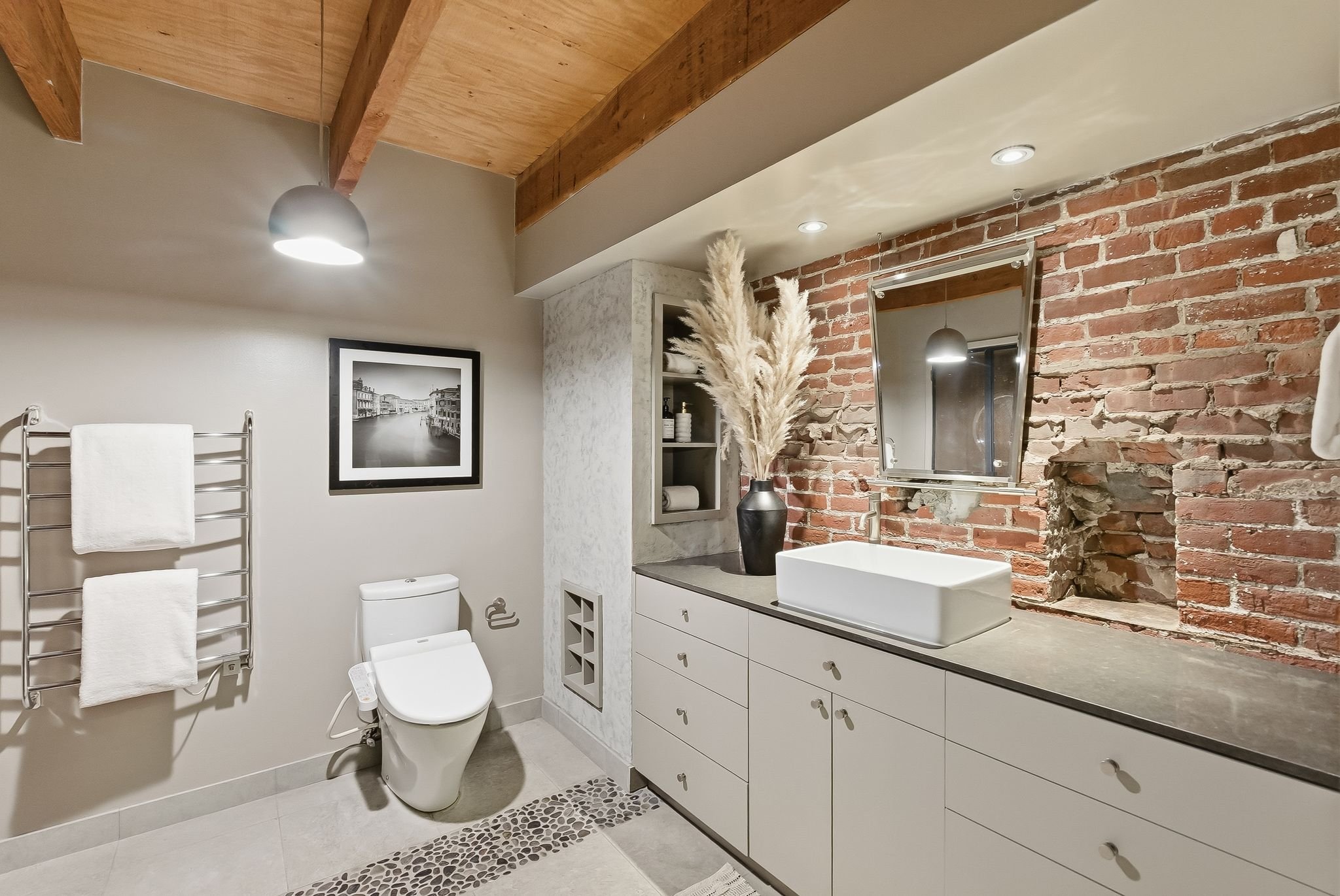 A modern bathroom with a white toilet, a white vanity with multiple drawers, a black vase with dried pampas grass, brick accent wall, a wall-mounted mirror, and a framed black and white cityscape photograph, illuminated by ceiling lights and a pendan