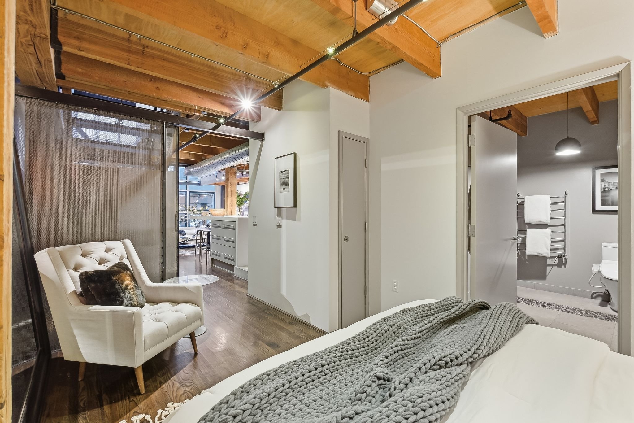 View of a modern apartment interior with wooden ceiling beams, a white armchair with a dog on it, a bed with a chunky knit blanket, and an open doorway leading to a bathroom with a towel rack and framed black-and-white pictures.