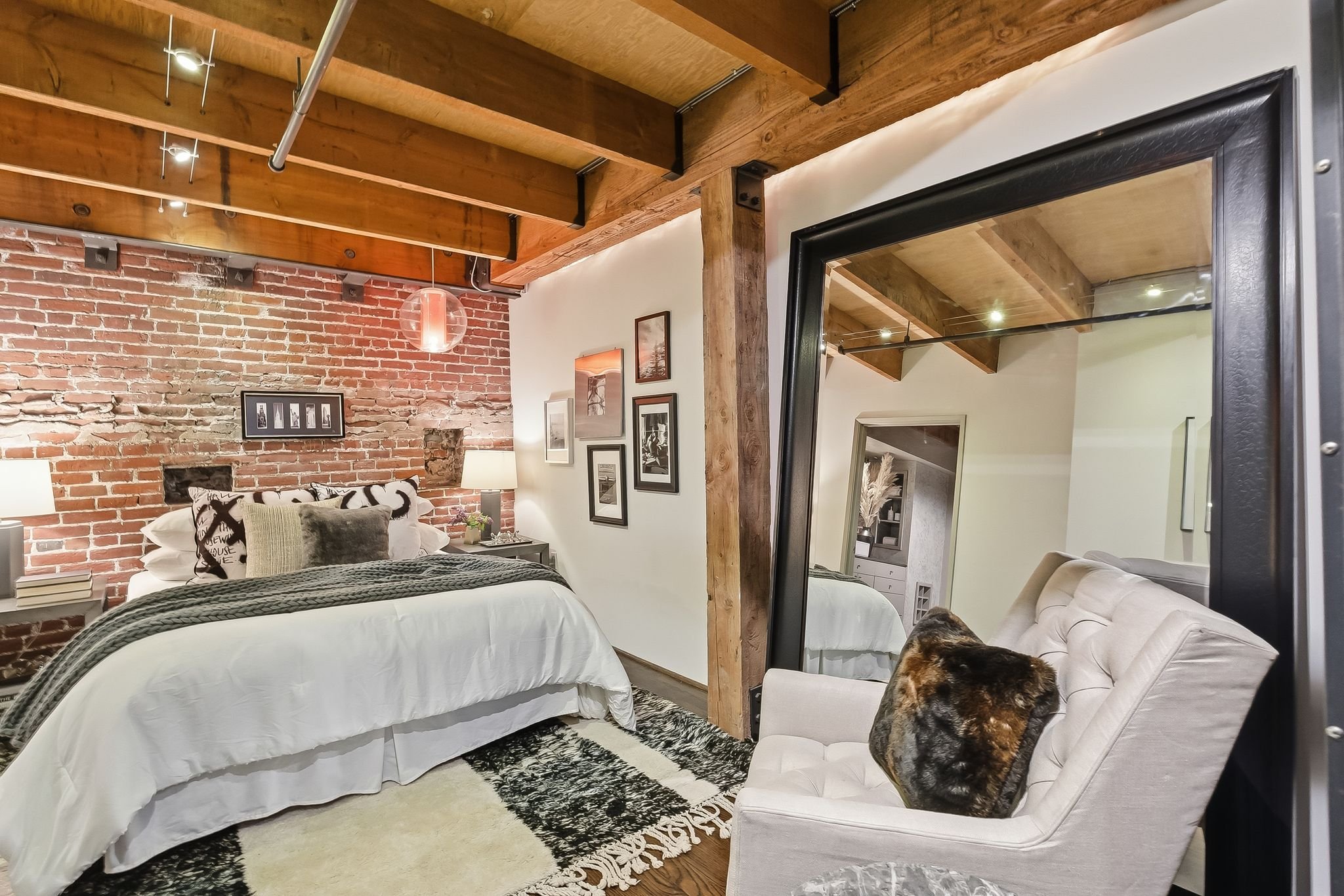 A bedroom with an exposed brick wall, wooden ceiling beams, and a large mirror reflecting part of the room. The bed has white bedding, multiple pillows, and a dark throw blanket. There is a loveseat with a plaid pillow and a dog resting on it. A fram