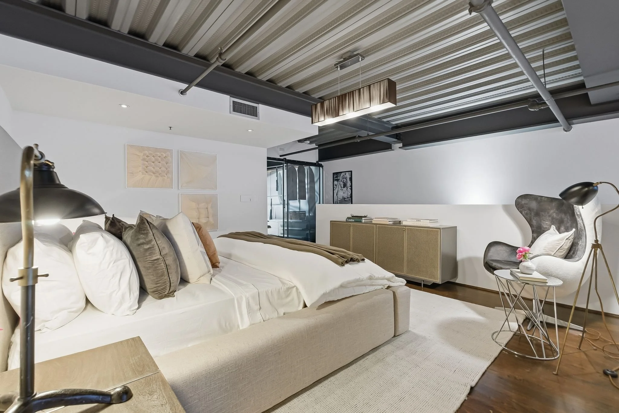 Modern living room with a low beige sofa, multiple pillows, a floor lamp, and a side table with books and a flower. The ceiling is industrial-style with exposed pipes and a wooden pendant light. Artwork hangs on the white wall, and there's a glass do