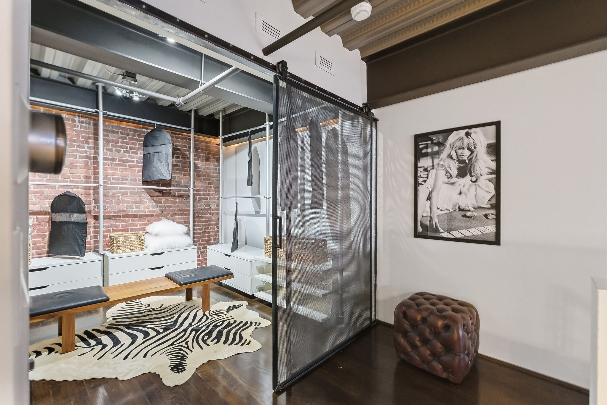 Interior view of a modern living space with a black metal and mesh room divider, exposed brick wall, black boxing bags, a wooden bench with black cushions, a zebra-patterned rug, and black and white artwork of a woman on the wall.