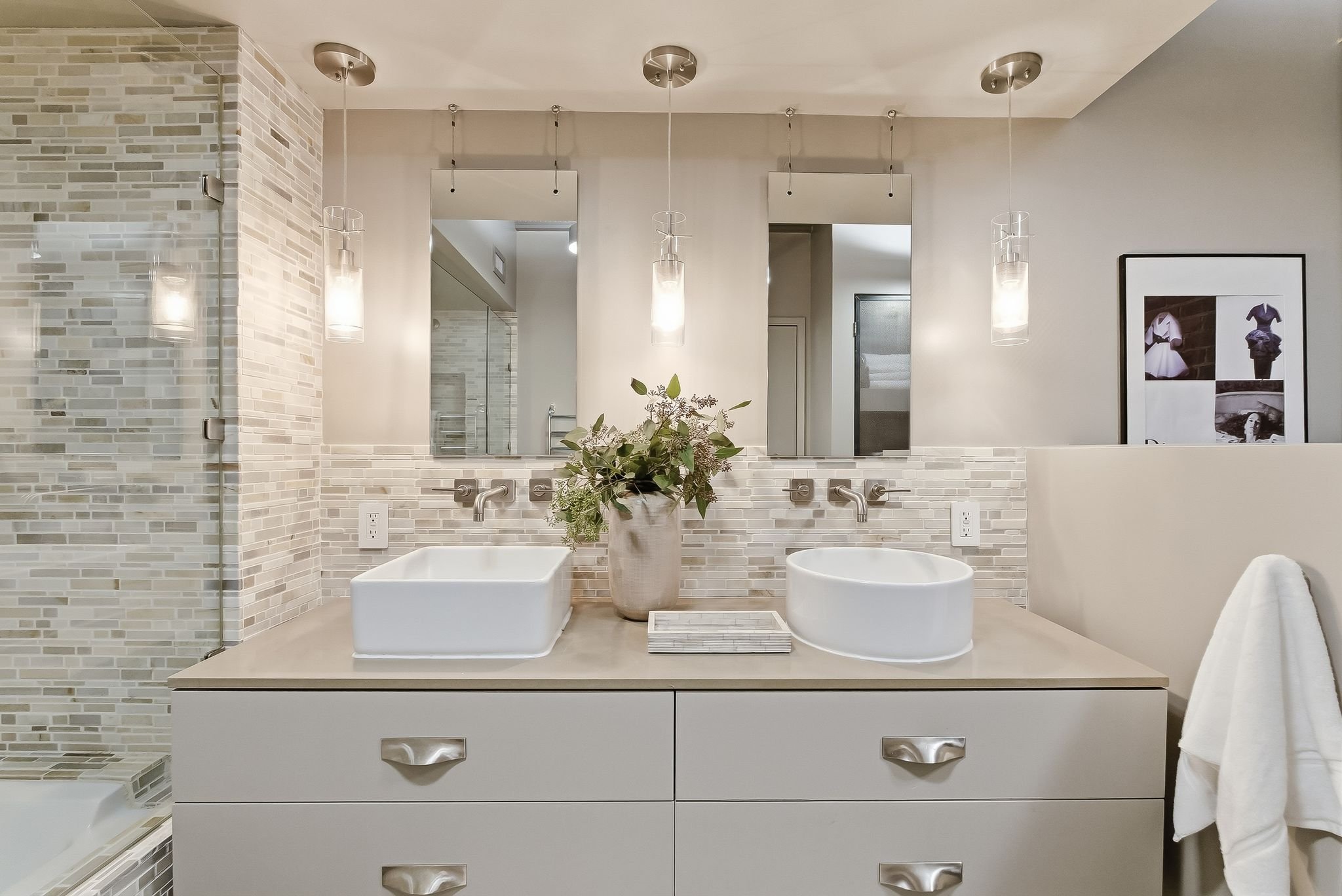Modern bathroom vanity with two vessel sinks, large mirrors, pendant lights, a vase with greenery, and tiled wall backsplash.
