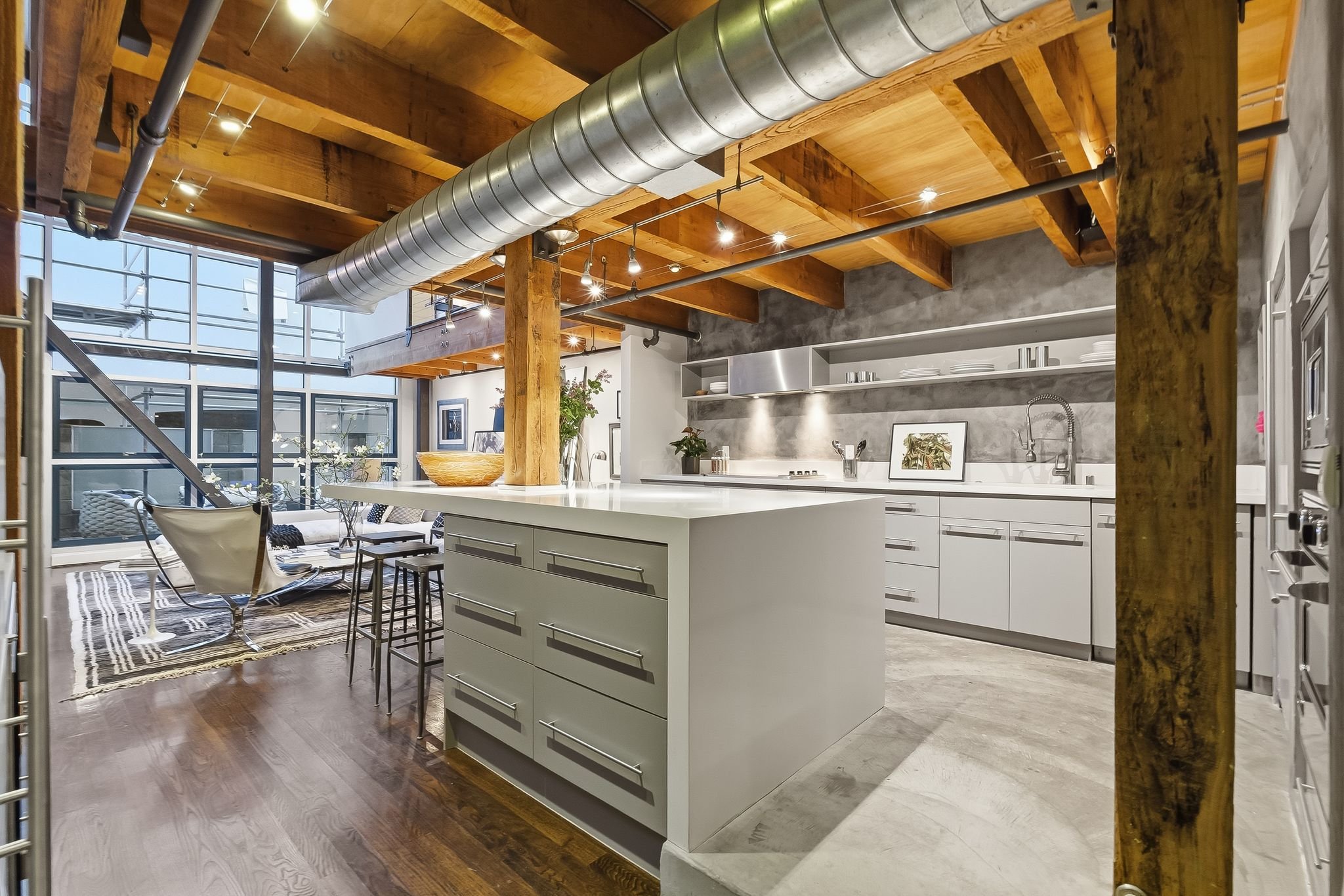 Modern kitchen with white cabinets, a large island, an exposed wooden beam ceiling, stainless steel appliances, and large windows in an urban loft setting.