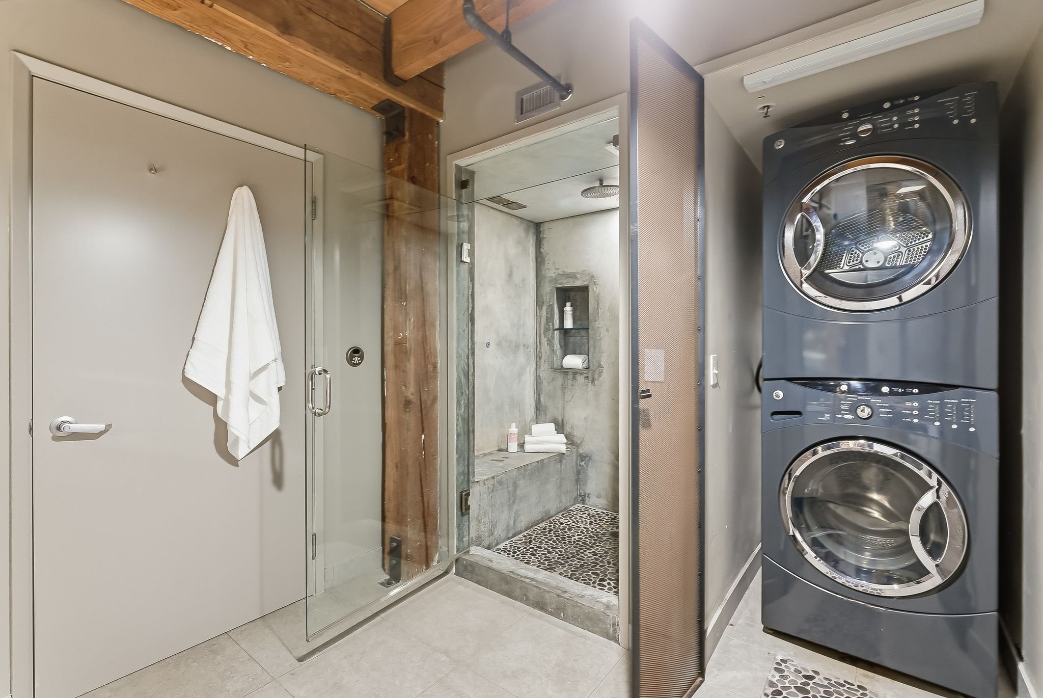 A modern laundry room with a stacked washer and dryer to the right, a glass-enclosed shower with wooden trim in the center, a gray door with a white towel hanging on a hook to the left, and a small shelf with toiletries inside the shower.