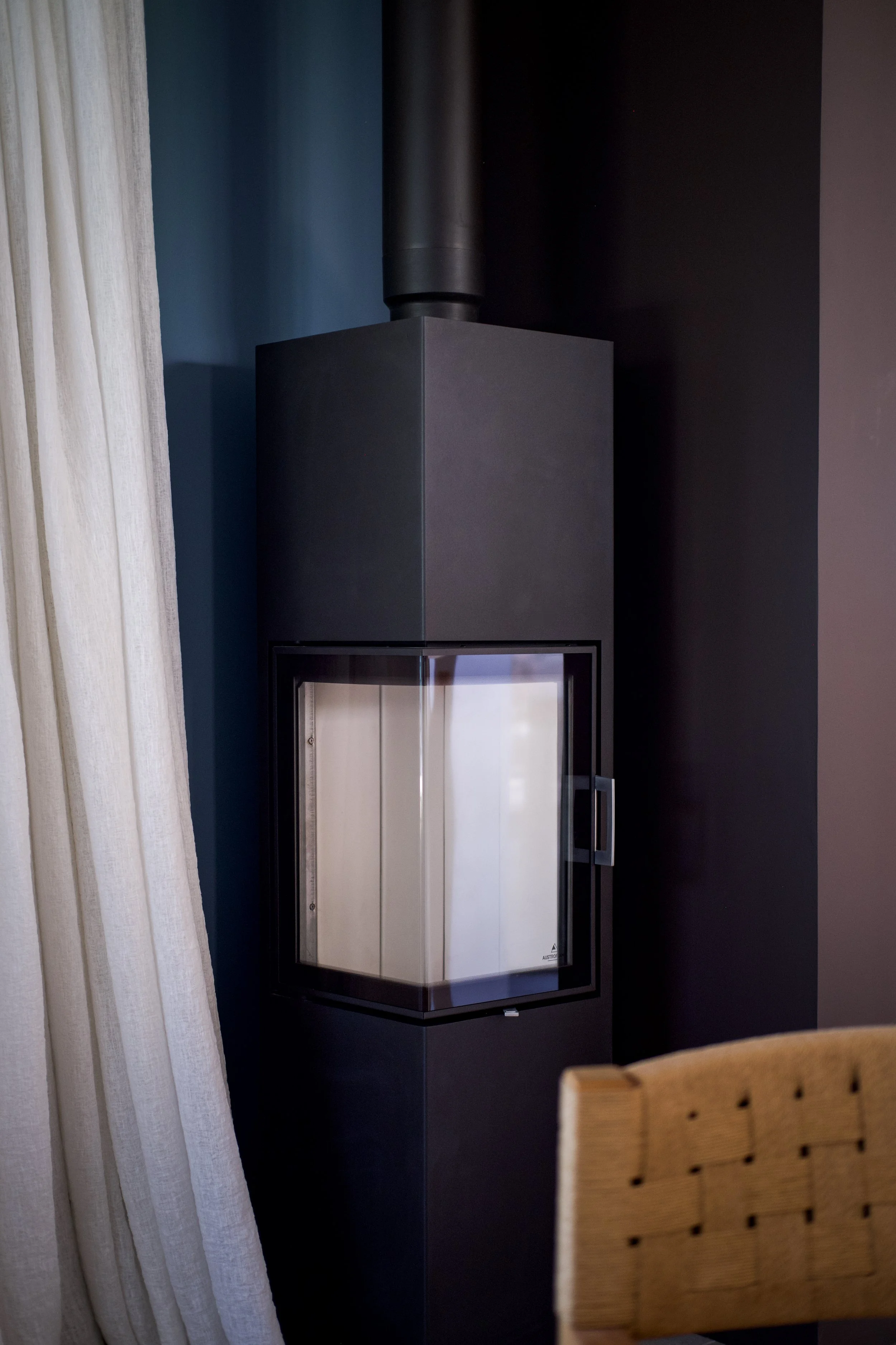 Photographie AirBnB Immobilier. Poêle à bois noir moderne dans une pièce avec un mur gris foncé, rideau beige et chaise en bois