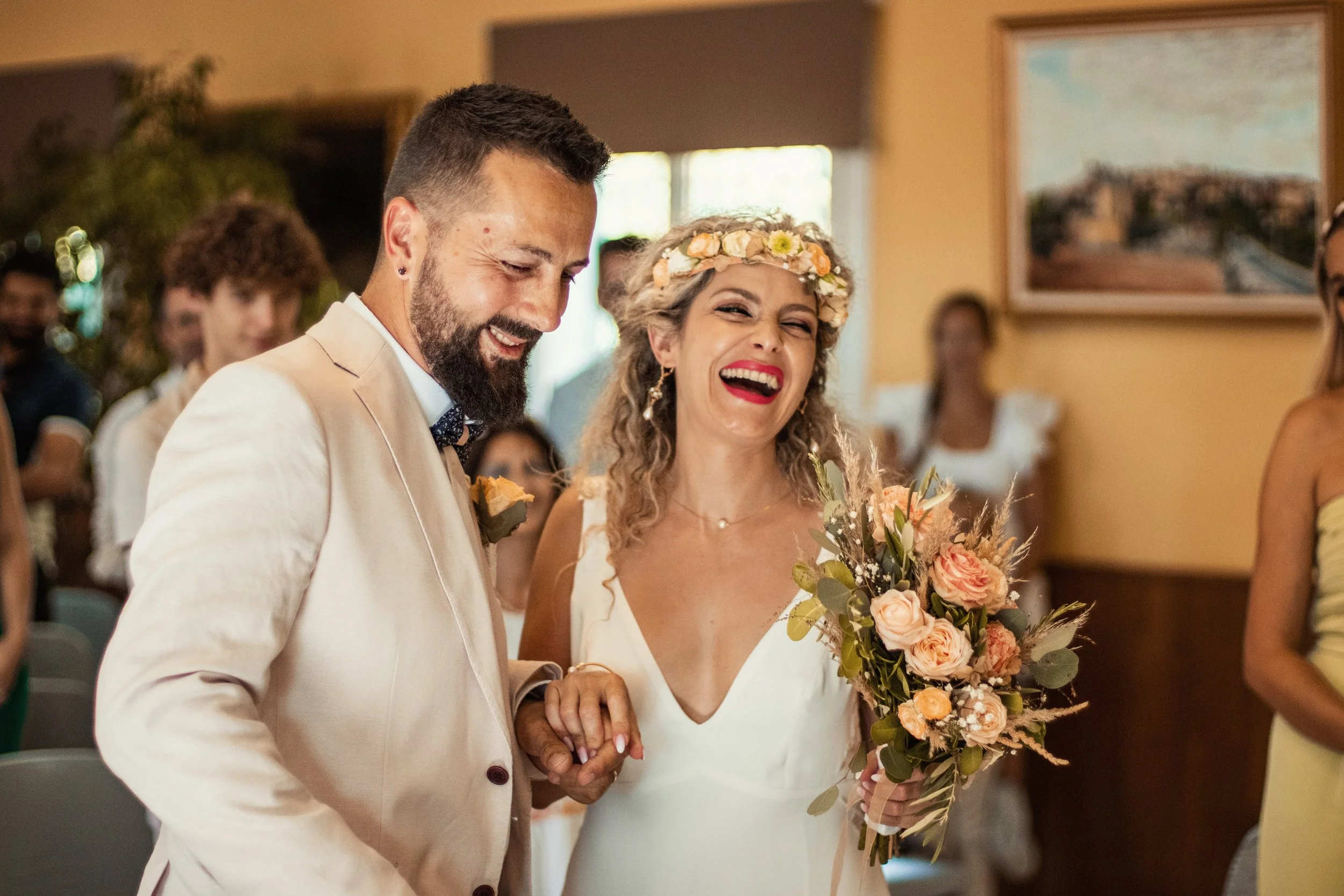 Photographe mariage - Un couple de mariés souriants lors de leur cérémonie de mariage, entourés d'invités, avec la mariée tenant un bouquet de fleurs et portant une couronne de fleurs, dans une salle décorée