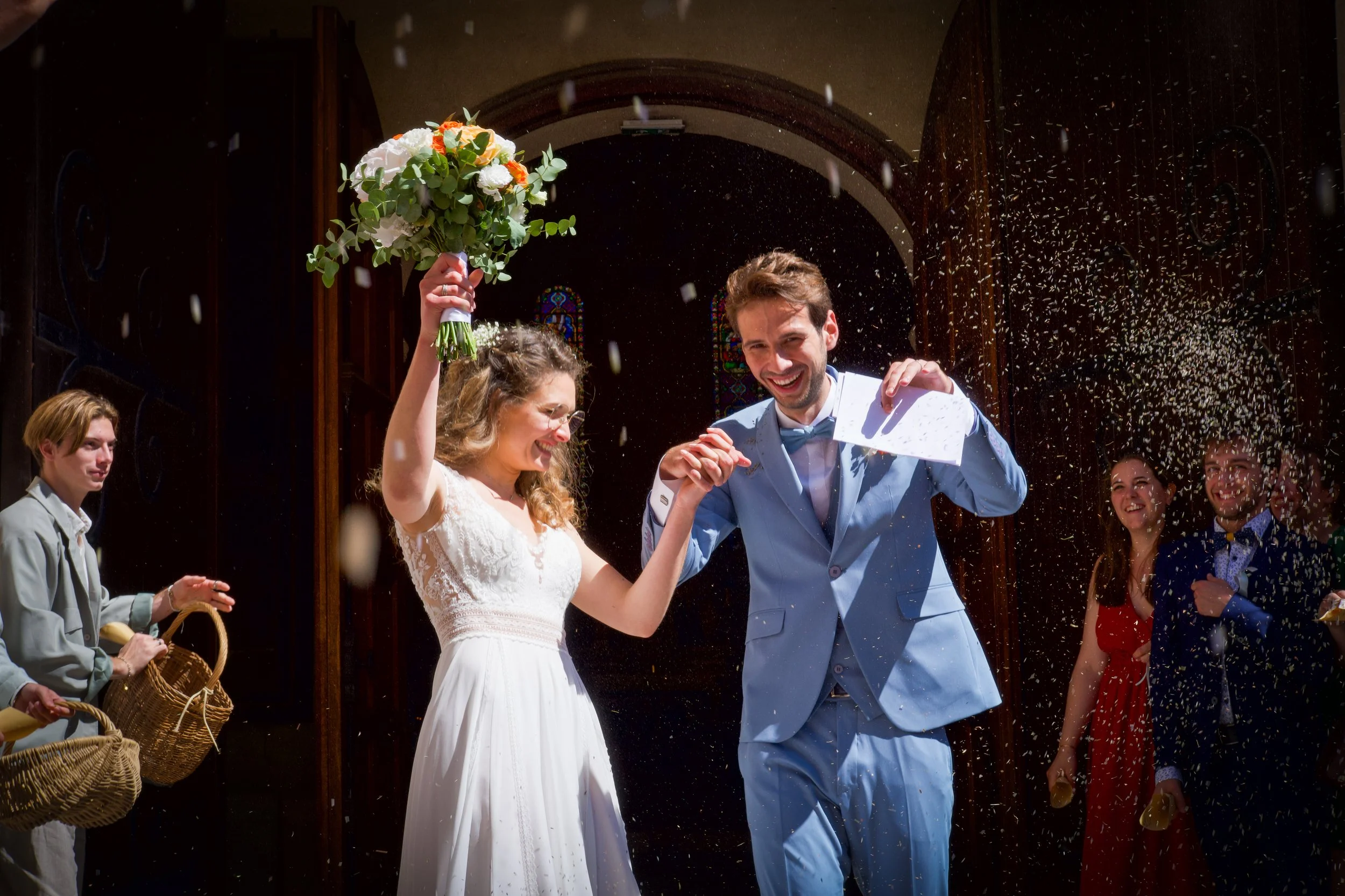 Photographe mariage - Un couple de mariés qui célèbre leur mariage en sortant de l'église, entourés d'invités. La mariée porte une robe blanche et tient un bouquet, le marié porte un costume bleu. Des invités dispersent des confettis autour d'eux.