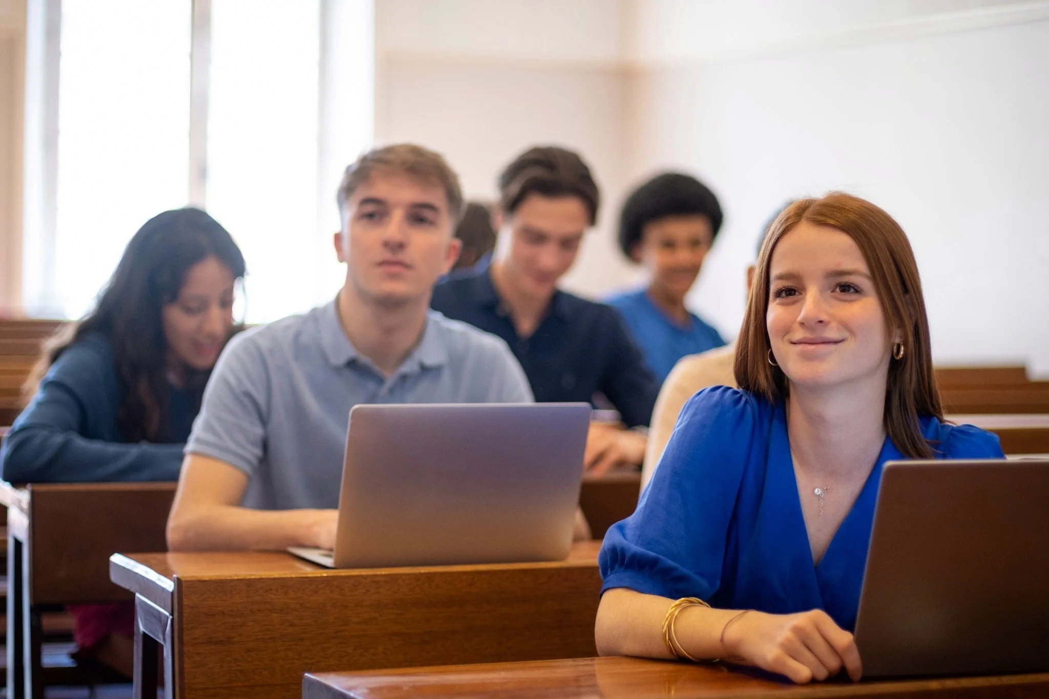 Photographe conférence - Étudiants assis en classe, certains utilisant des ordinateurs portables, une jeune femme souriante en premier plan, une salle de classe lumineuse.