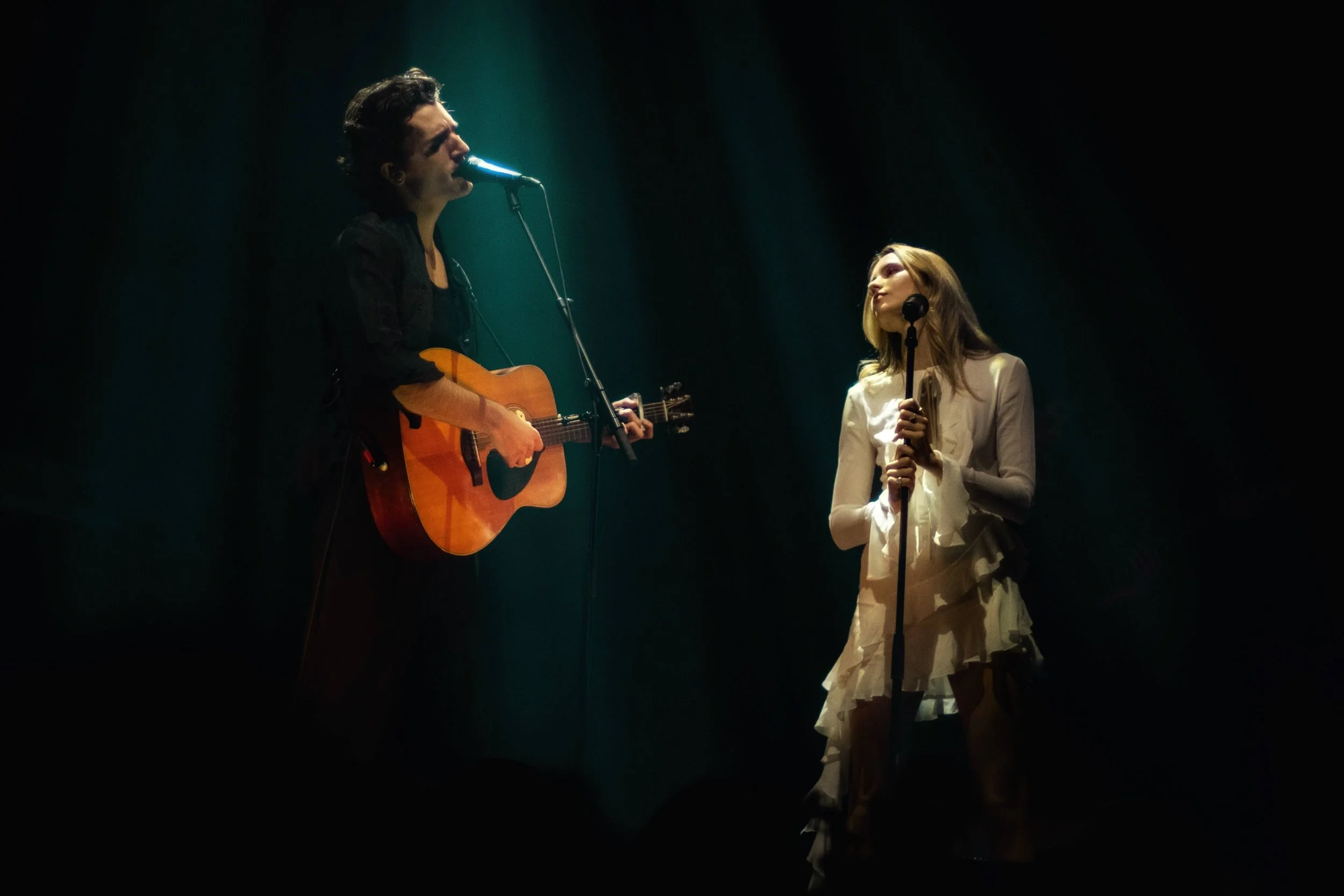 Photographe concert - Un homme joue de la guitare et chante dans un micro, une femme chante avec un microphone sur une scène sombre.
