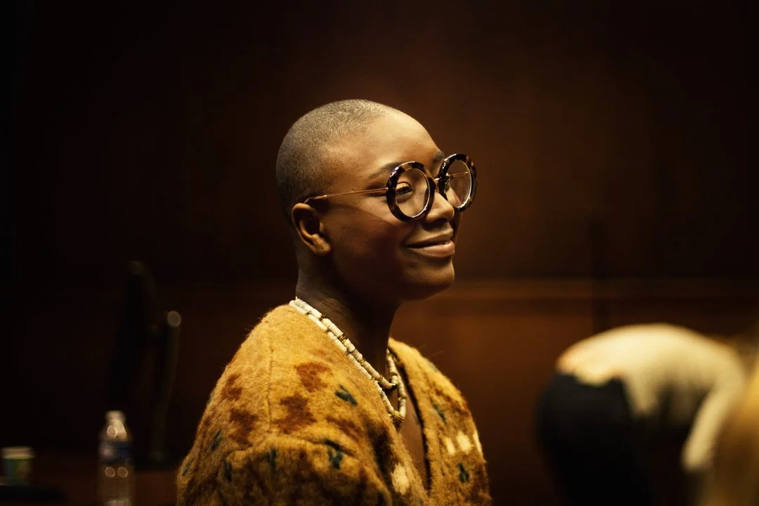 Photographie événement - Une femme avec une tête rasée, portant de grosses lunettes rondes et un manteau brun, sourit dans un environnement intérieur sombre.