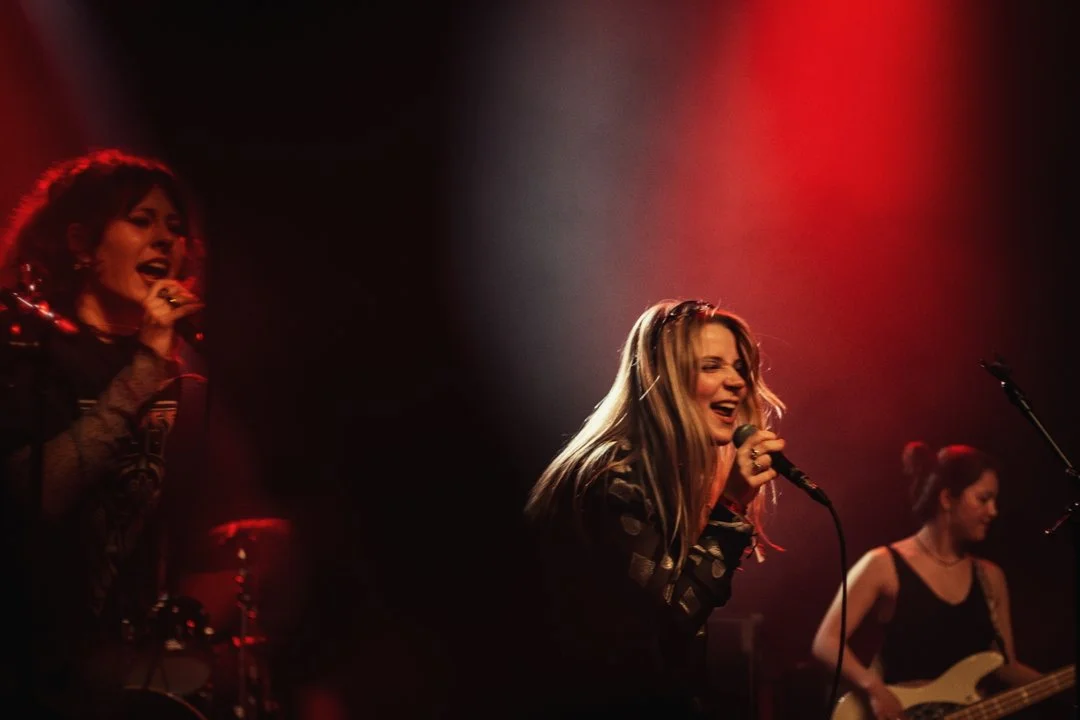Photographe concert - Groupe de femmes chanteuses sur scène lors d'une performance musicale, éclairage rouge et clair sur elles.