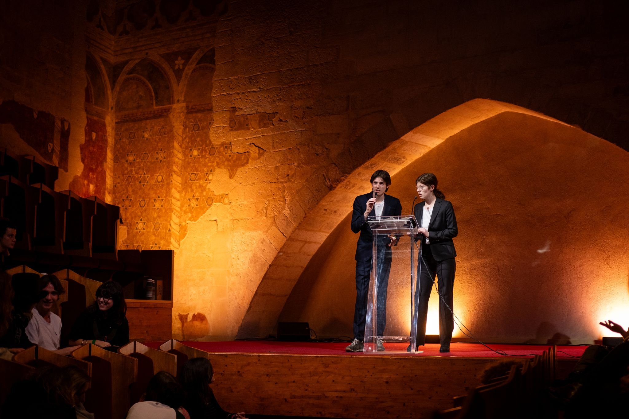 Photographe événement - Deux personnes en costume noir parlent à un micro derrière un pupitre transparent sur une scène avec un fond en pierre et lumière chaleureuse. Audience assise en bas.